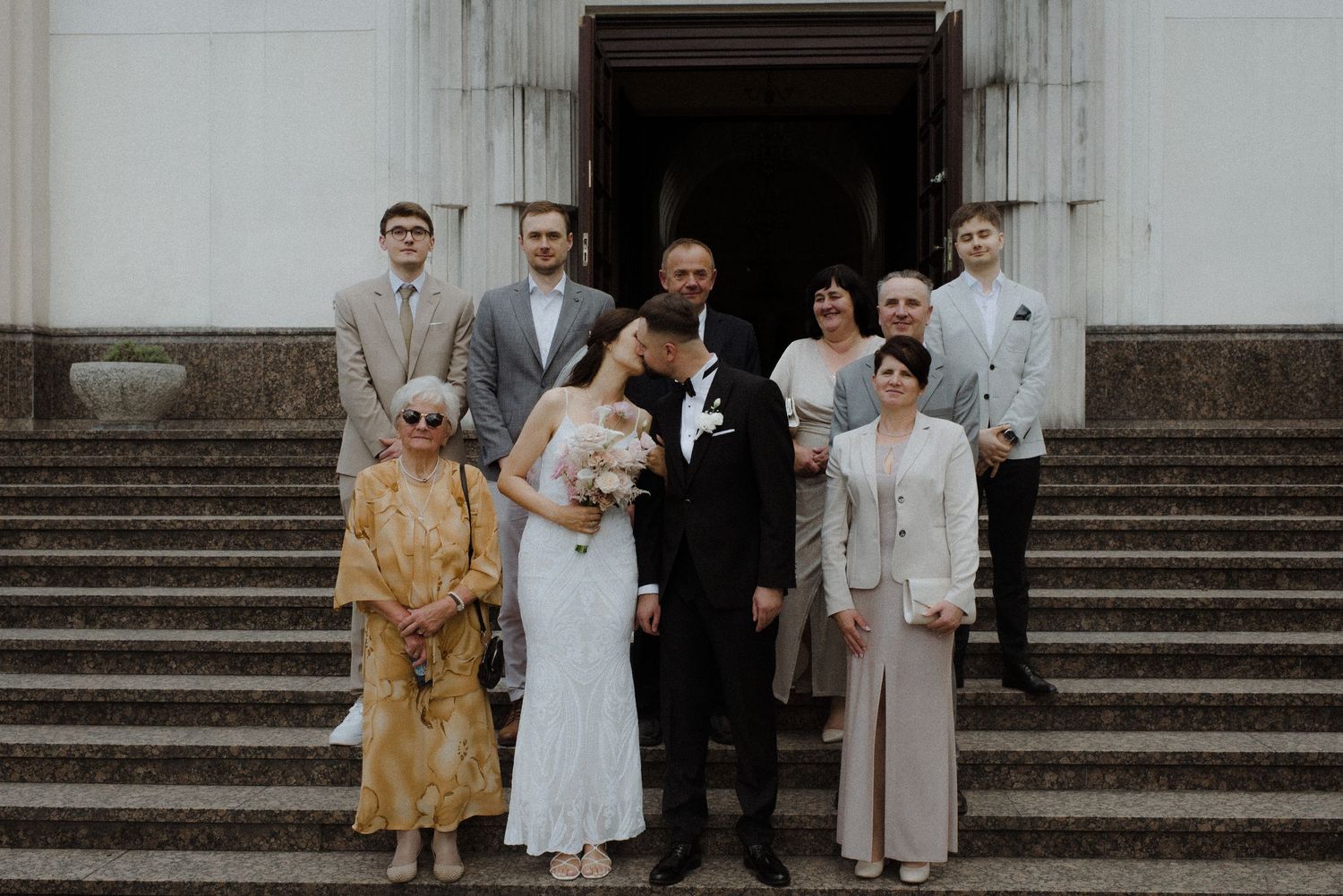 Wedding party group poses on elegant stone steps outside historic building.