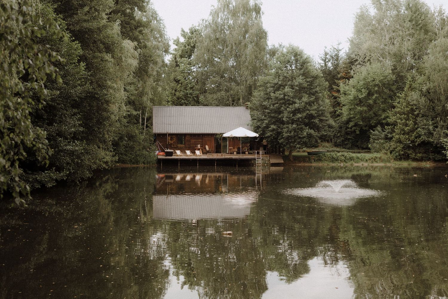 A wooden cabin reflects perfectly in a serene lake with a fountain spray in the center.