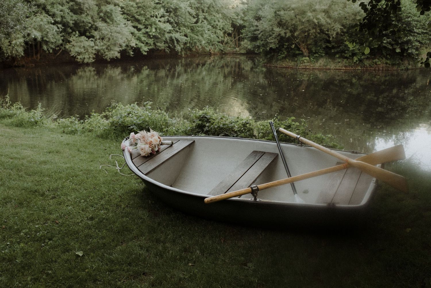 An empty wooden rowboat rests on the grassy shore of a calm lake.