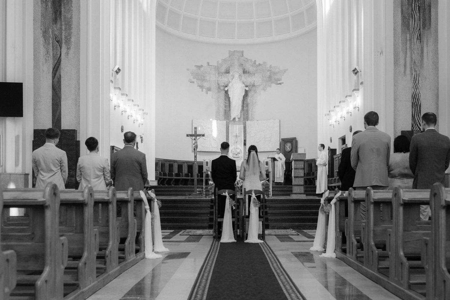 Black and white photograph showing interior of a church sanctuary with rows of empty wooden pews and vaulted ceiling.