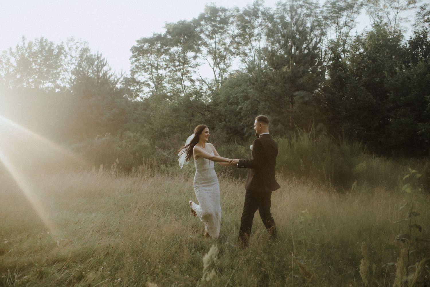 Newlyweds walk together through a sunlit meadow surrounded by lush greenery.