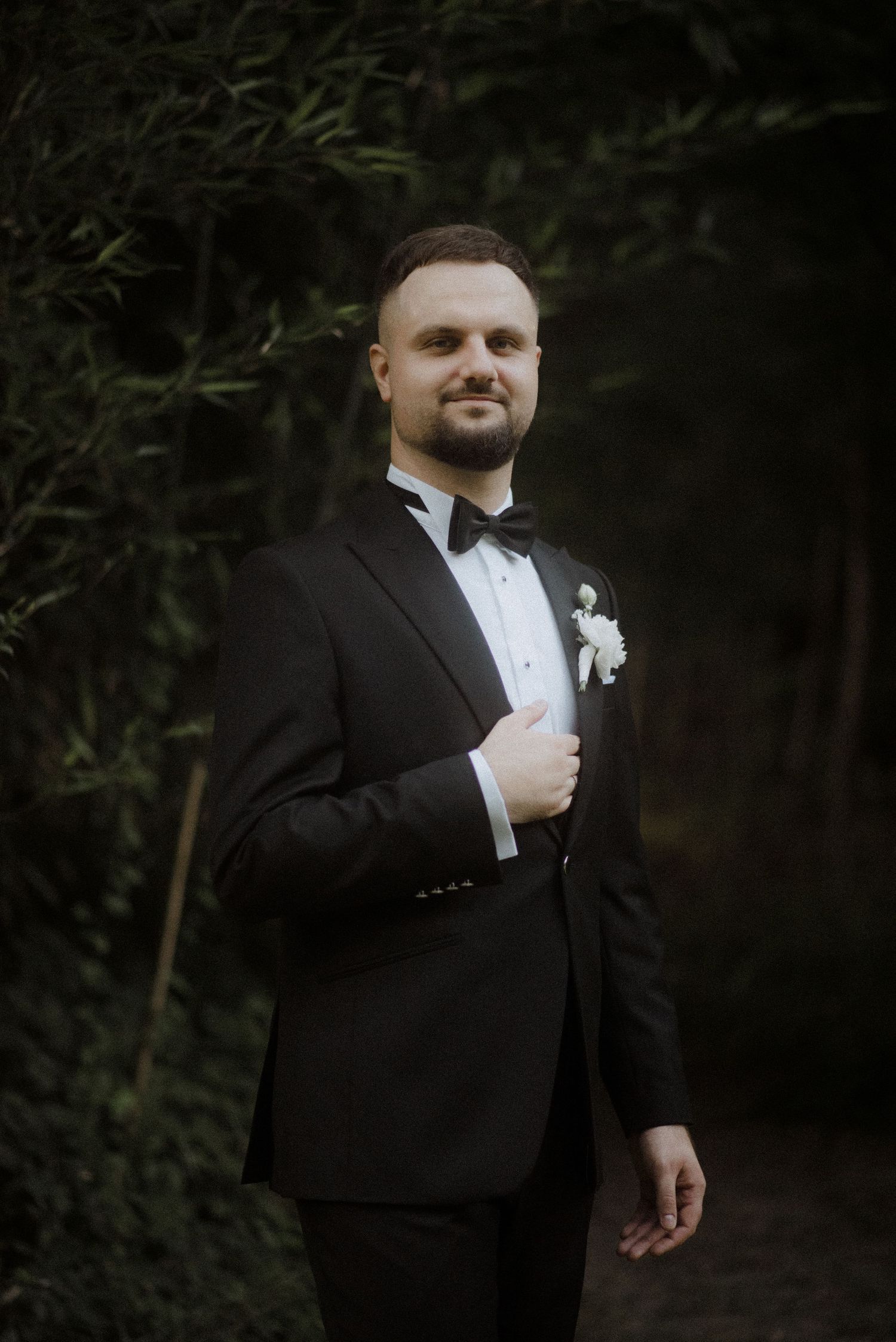 Groom in a classic black tuxedo and bowtie adjusts his boutonniere outdoors.
