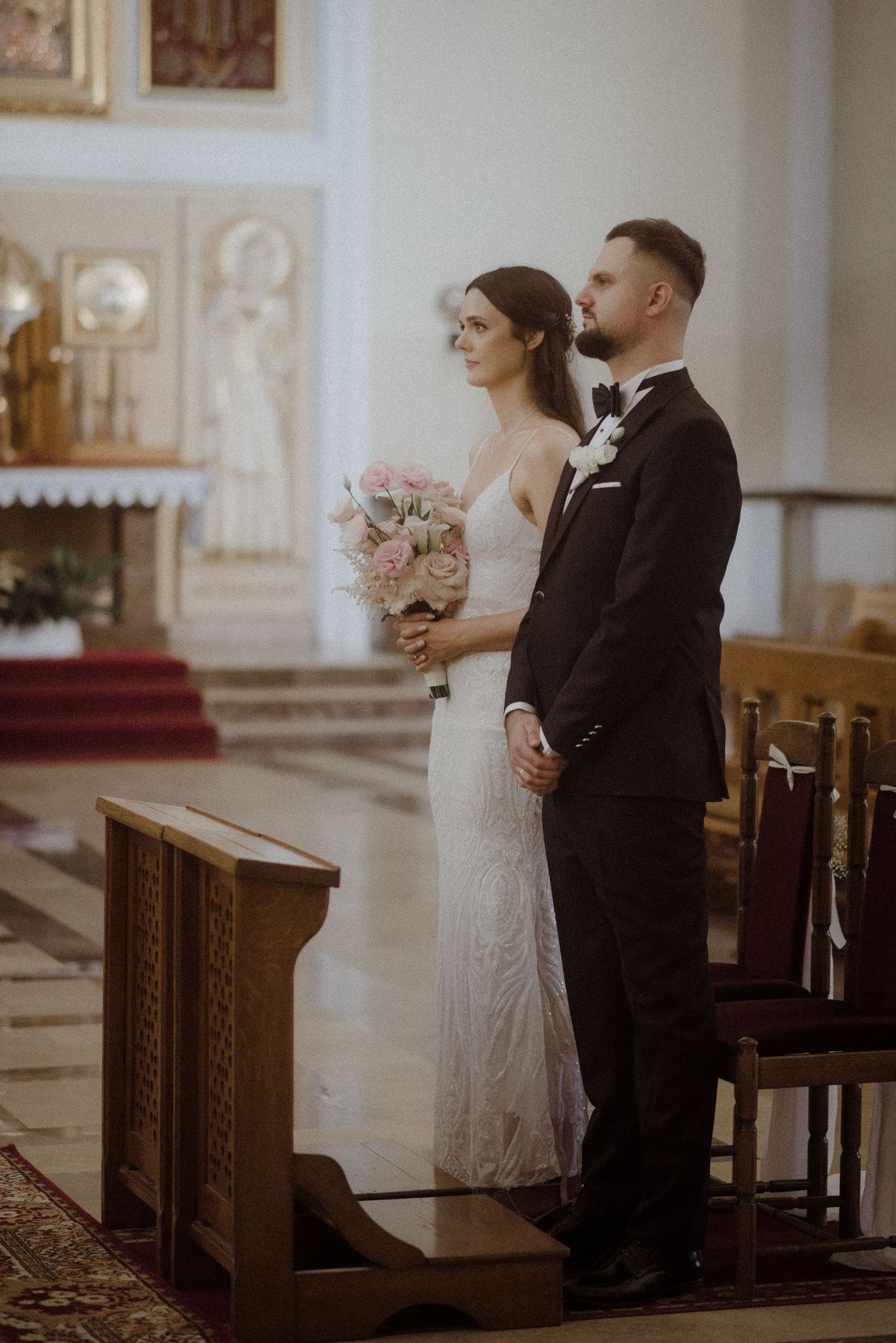 A couple stands together in a traditional church during their wedding ceremony, surrounded by wooden pews and ornate decor.