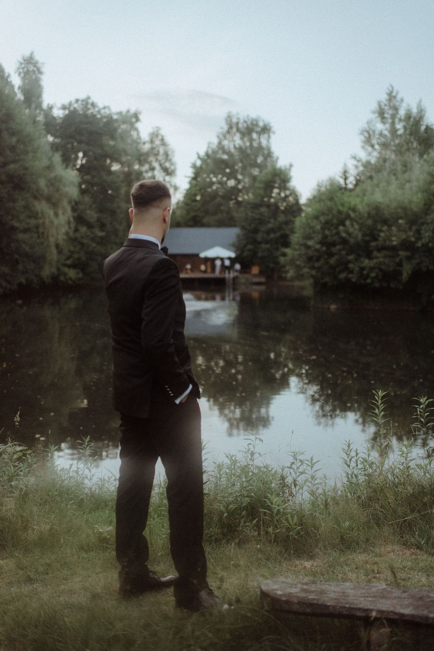 Two people in formal attire standing by a tranquil lake with reflections on the water.