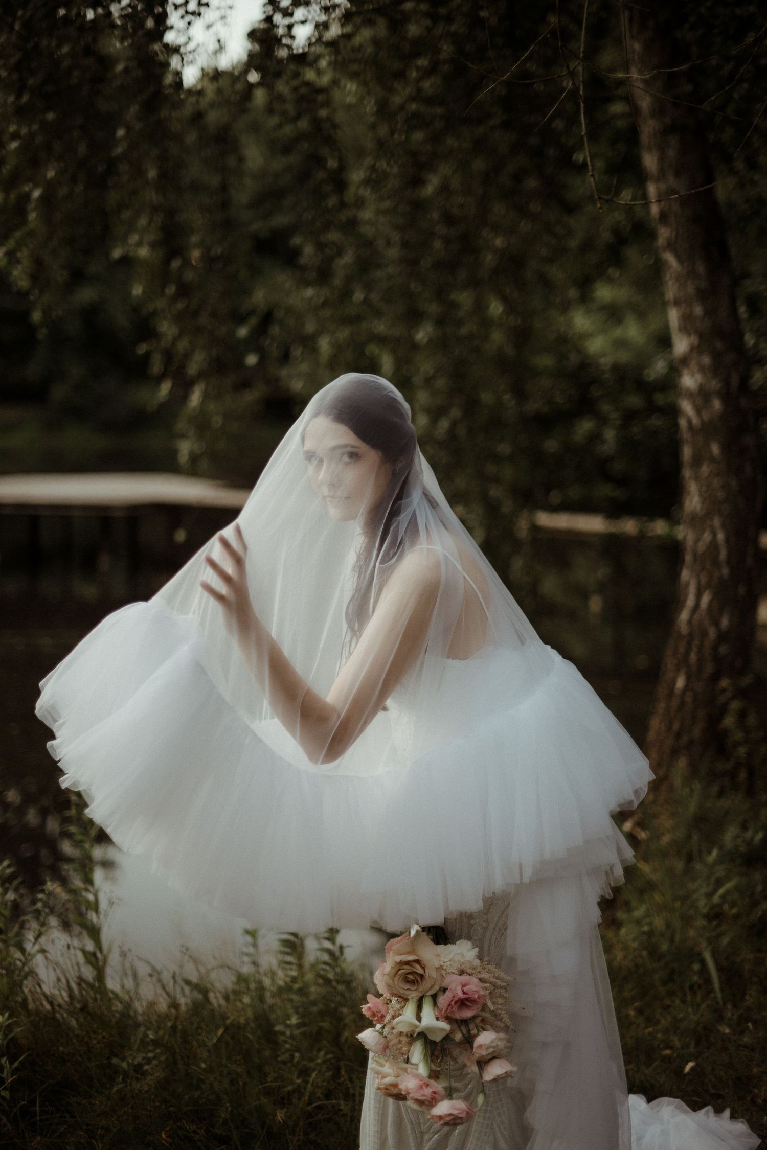 Ethereal bride in flowing tulle dress and veil poses in forest setting.