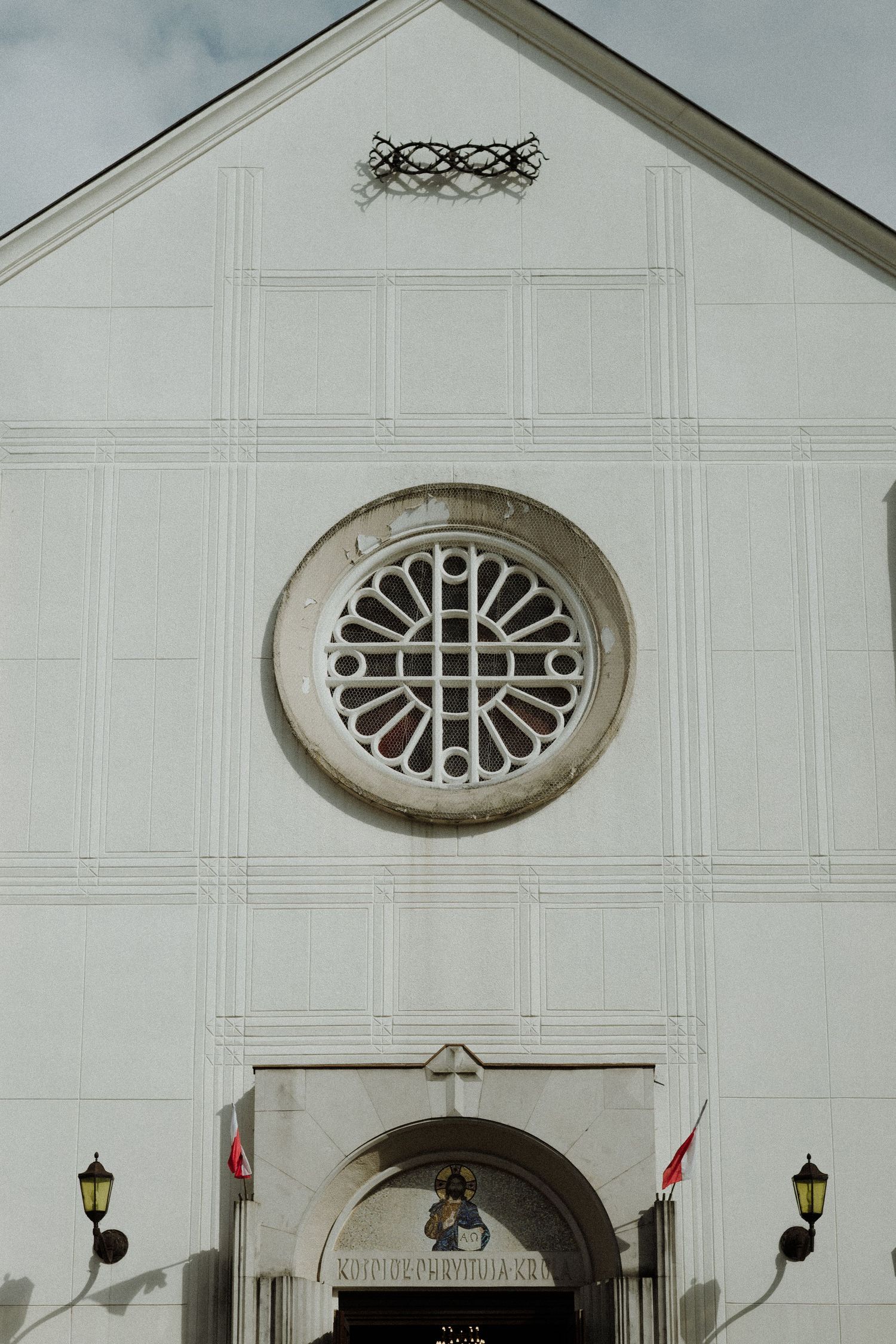 White church facade with ornate circular window and arched entrance featuring decorative trim and mounted lanterns.