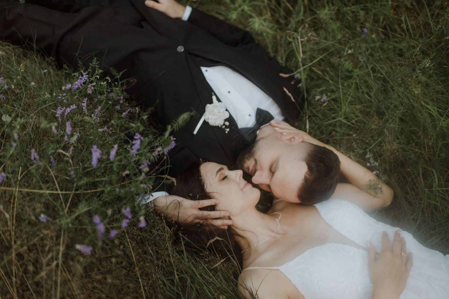 A couple lying in tall grass surrounded by purple wildflowers during an outdoor photoshoot.