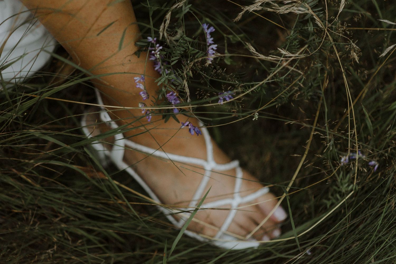 White heels with purple flowers sit nestled in tall grass and wildflowers creating a romantic outdoor setting.