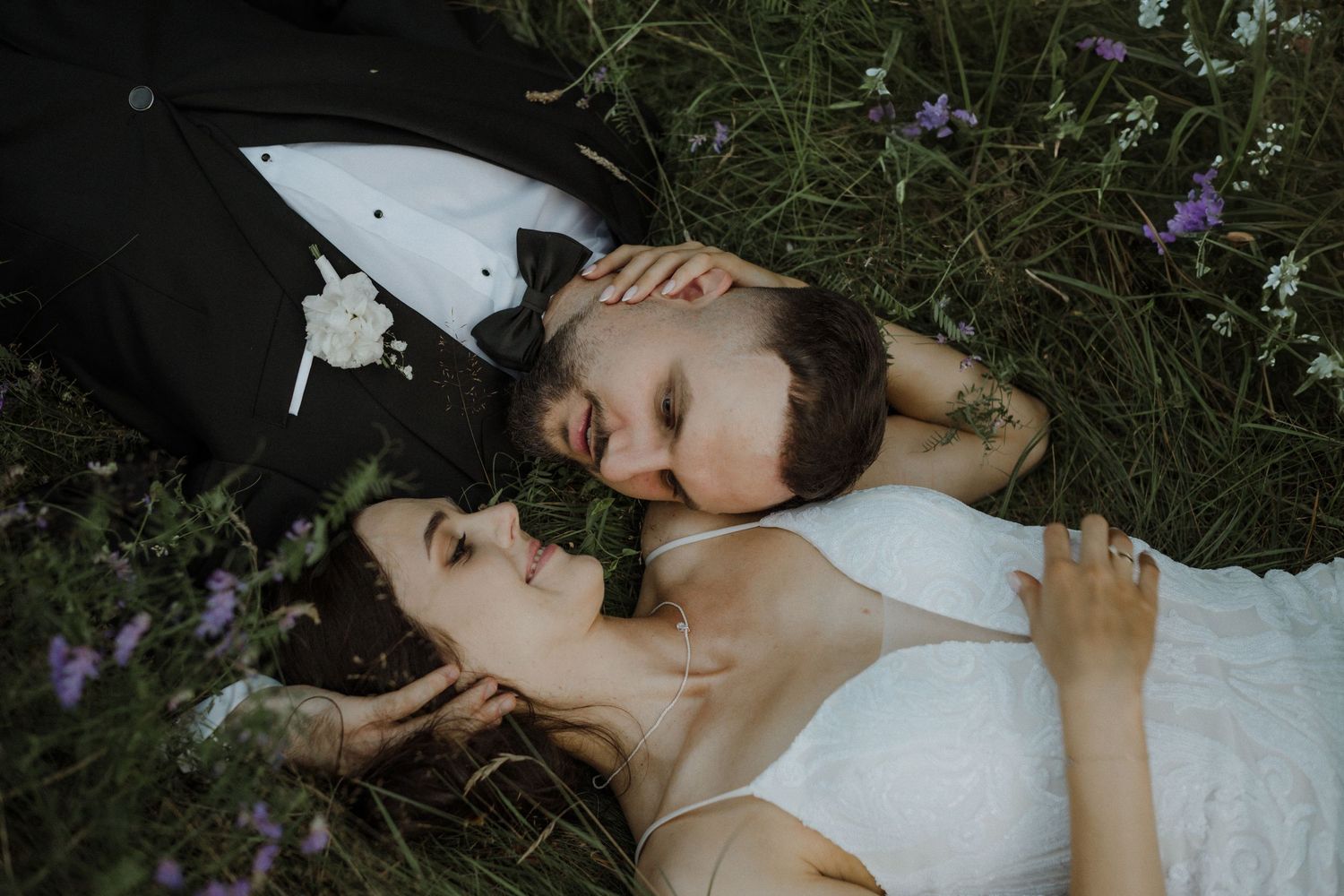 Couple lying in grass surrounded by wildflowers in formal attire.