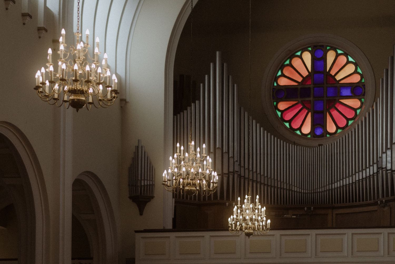 Ornate church interior featuring crystal chandeliers, organ pipes, and a colorful stained glass rose window.