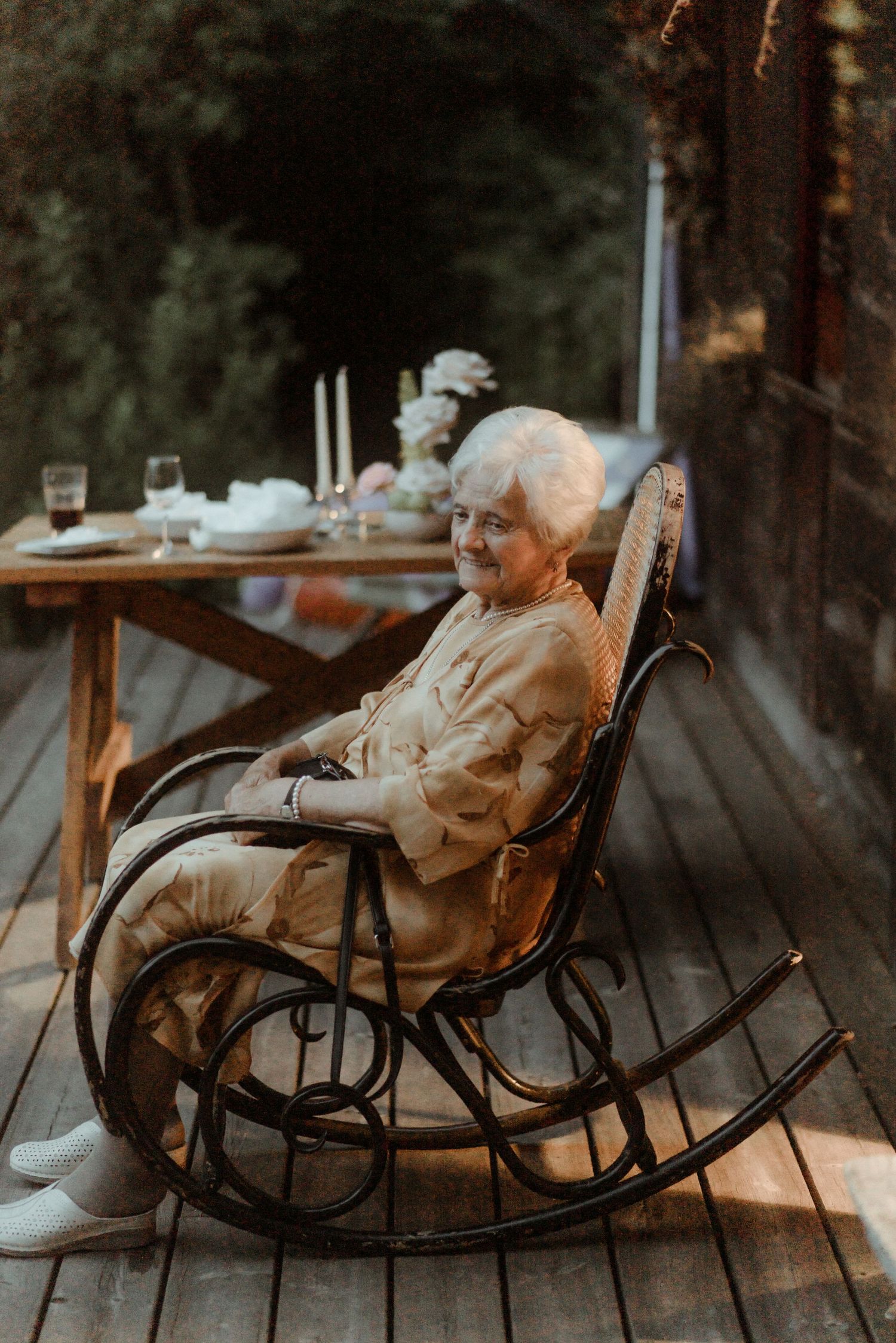An elderly woman relaxes in a vintage rocking chair on a wooden porch with a table setting behind her.