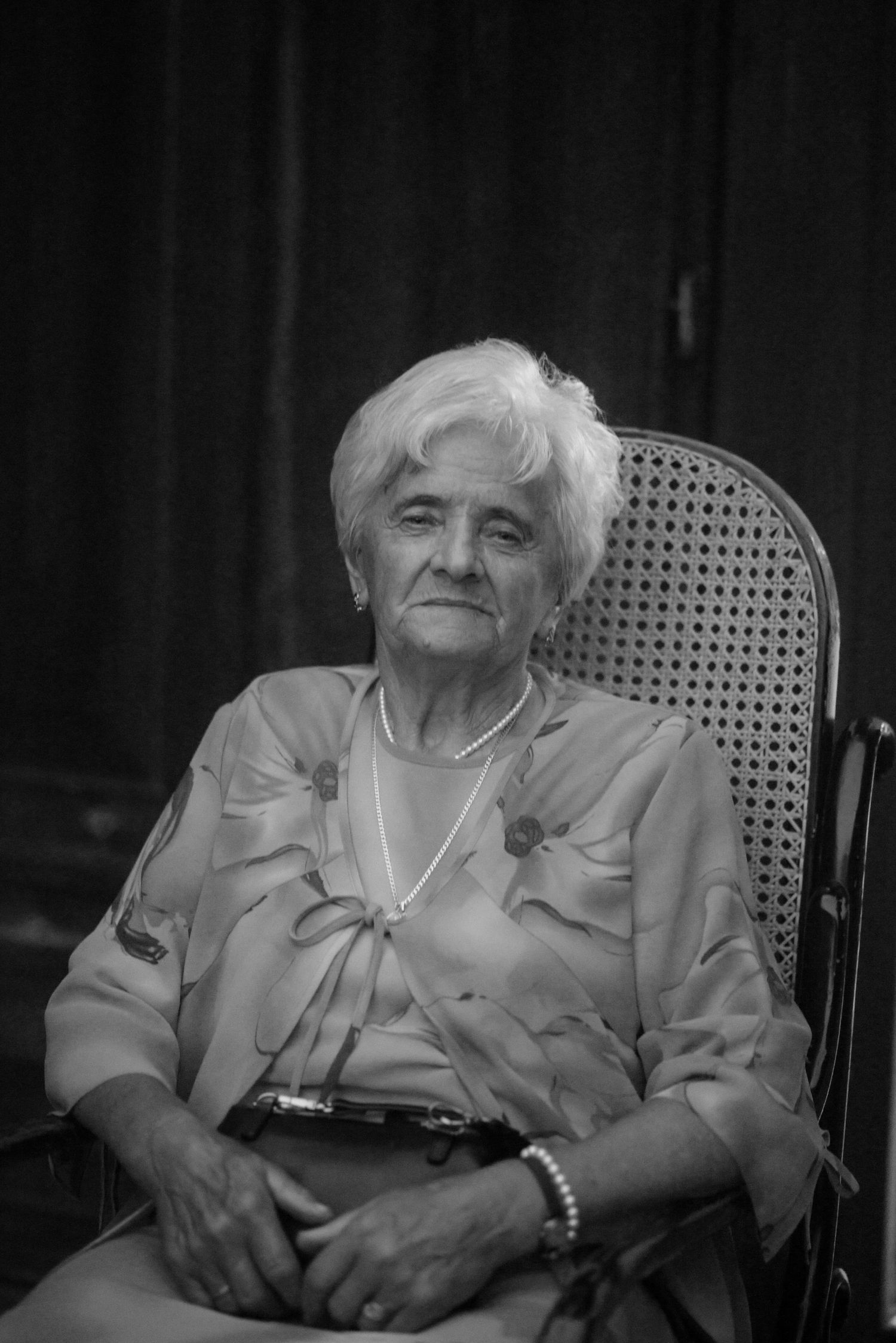 An elderly woman sits in a cane chair, wearing a light-colored outfit in this black and white portrait.