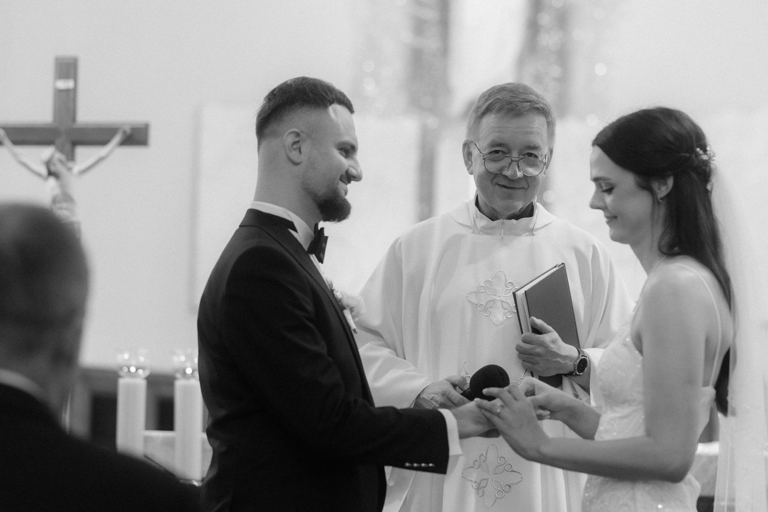 Black and white wedding ceremony photo showing couple and priest in church during exchange of rings.