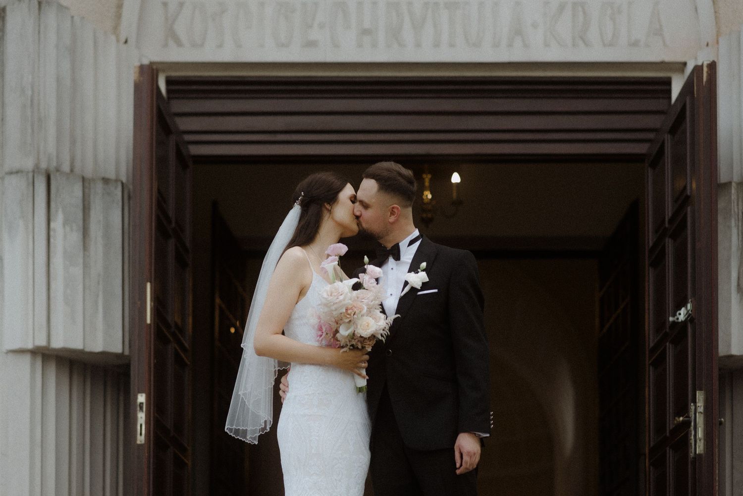 A romantic wedding scene showing a couple in elegant attire standing in front of rustic wooden doors.