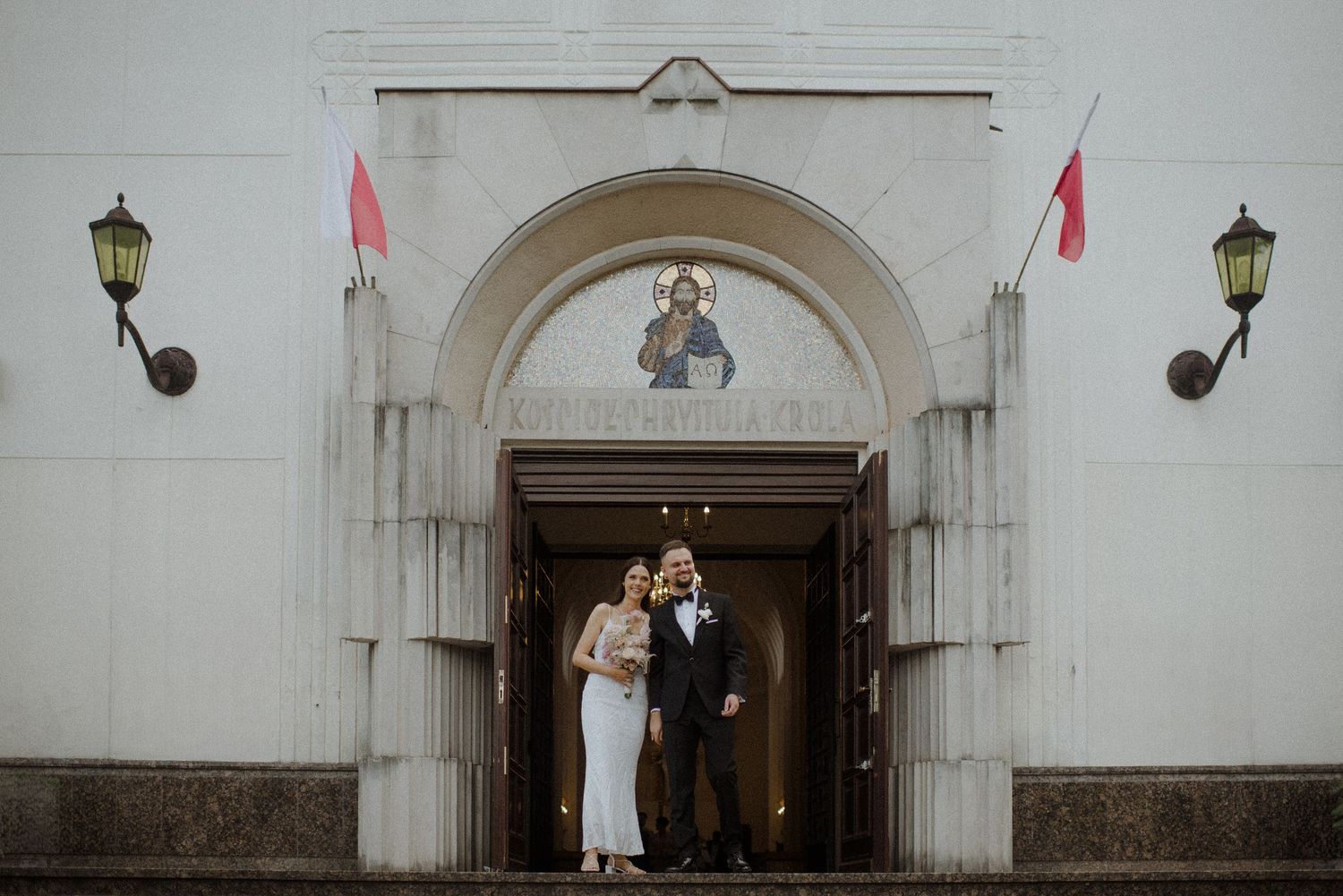 A bride and groom walking through an elegant white archway decorated with pink ribbons and flowers.