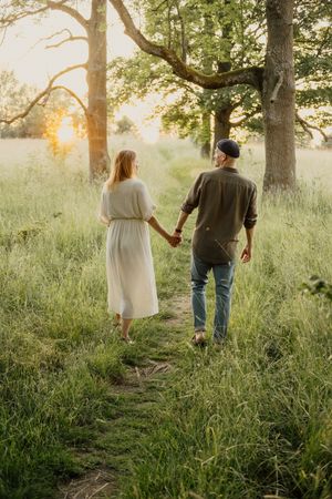 Couple holding hands walking on a sunlit path through lush green meadow at golden hour.