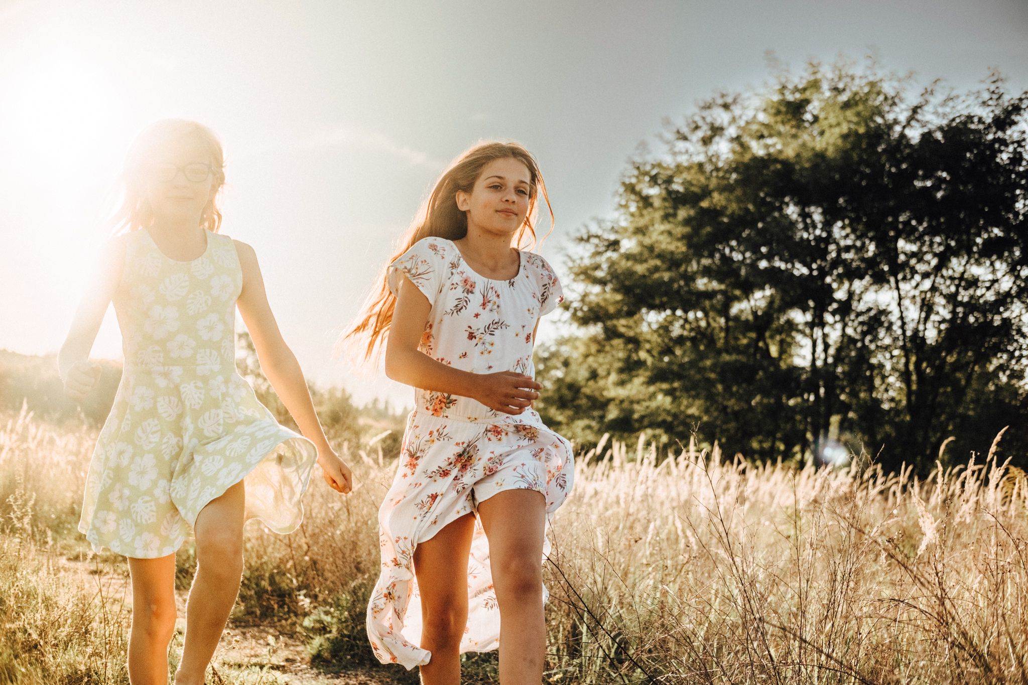 Girls running through a sun-lit field of tall grass during summer.