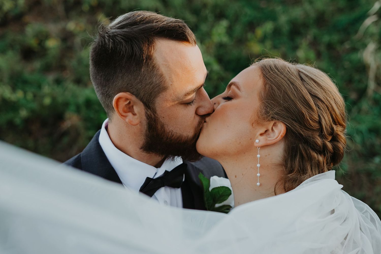 A romantic wedding moment captured as a couple shares a kiss outdoors with greenery in the background.