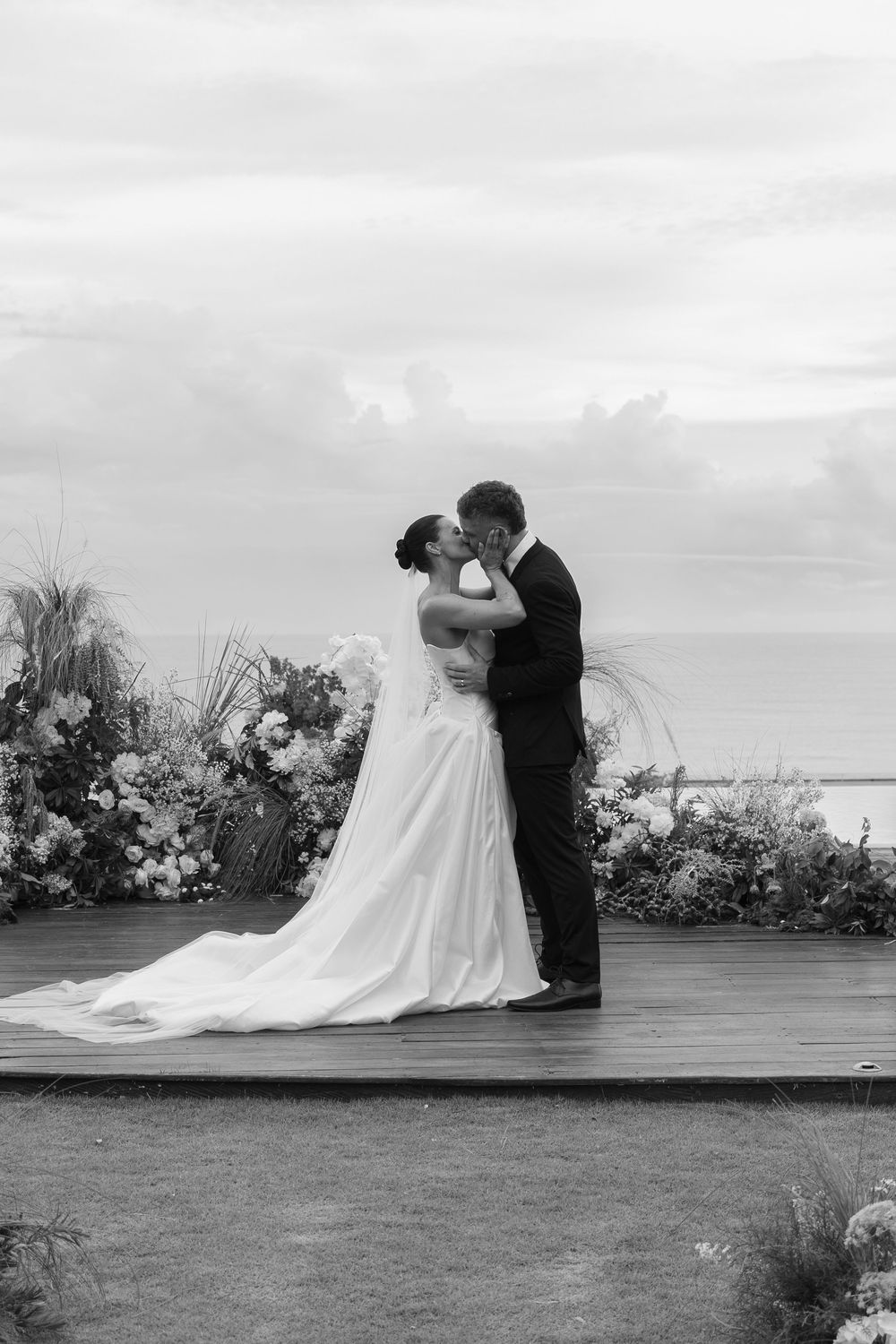 Black and white panoramic wedding photo showing tender moment between newlyweds on scenic oceanfront lawn with flowers.