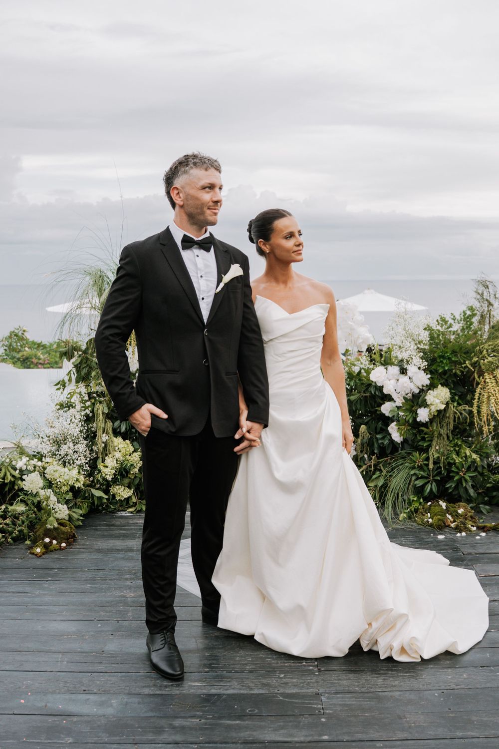 A couple in formal wedding attire walks together on a deck with mountain and floral backdrop.