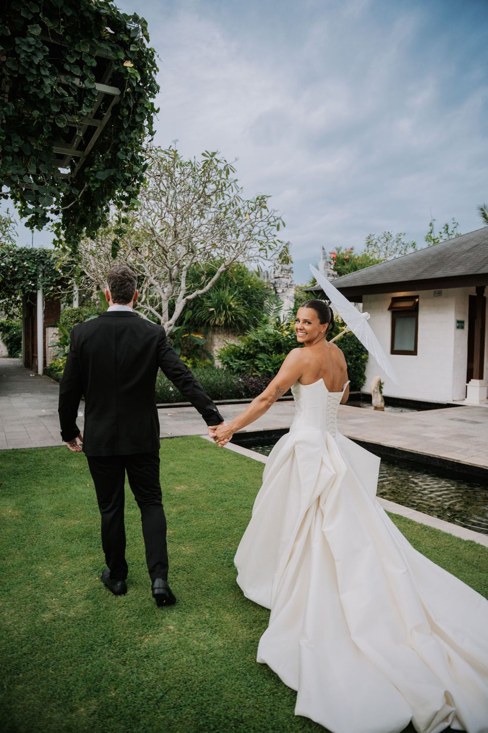 A wedding couple strolls hand in hand across a lush green lawn on an overcast day.