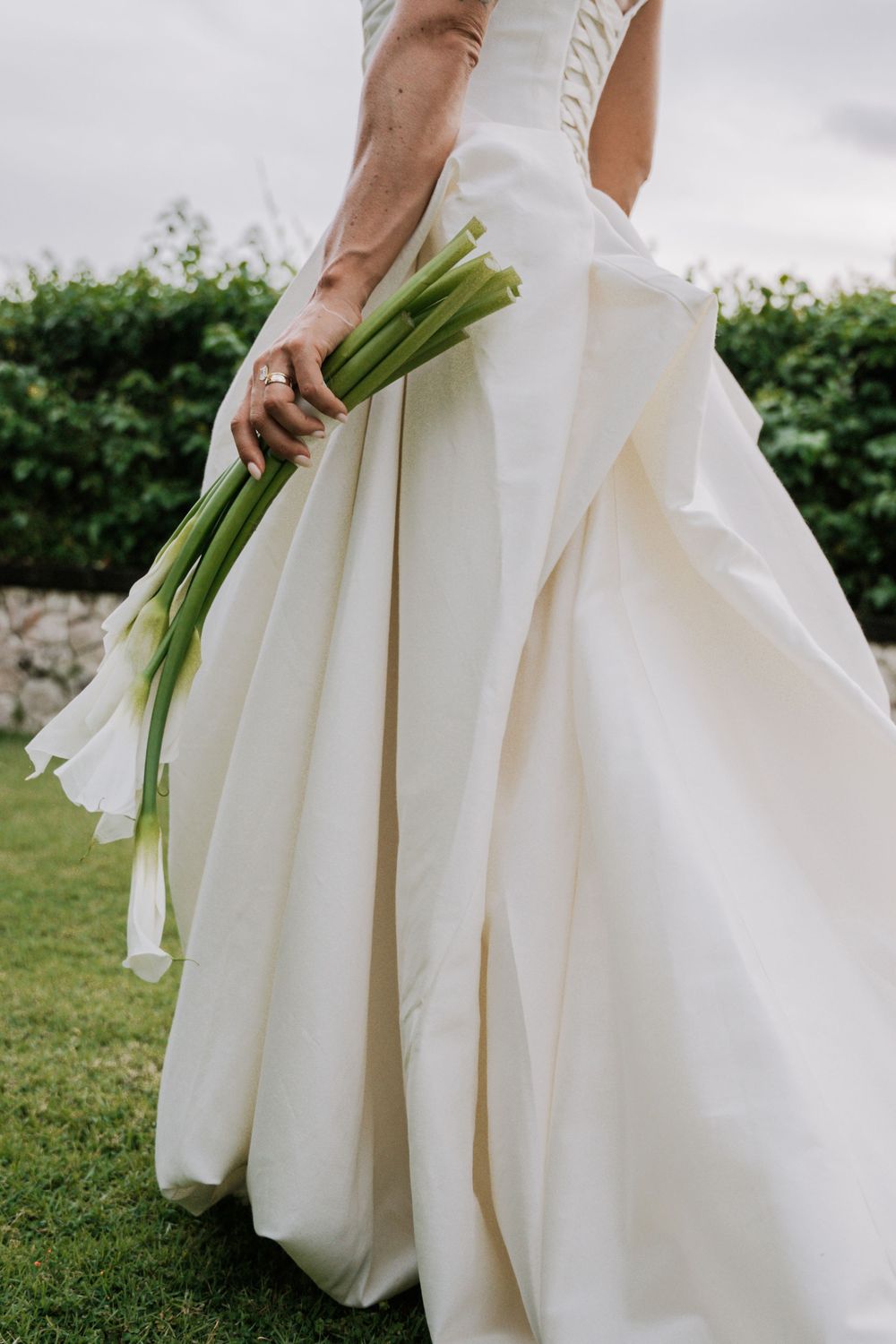 Wedding dress detail showing flowing white fabric and hands holding a green floral bouquet in an outdoor garden setting.