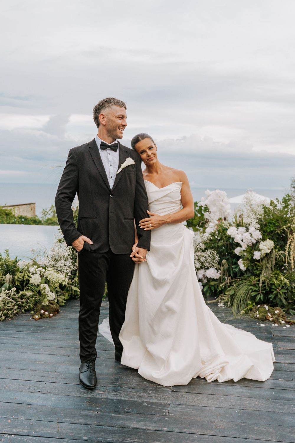 Newlywed couple sharing a romantic moment on outdoor deck surrounded by white floral arrangements under cloudy sky.