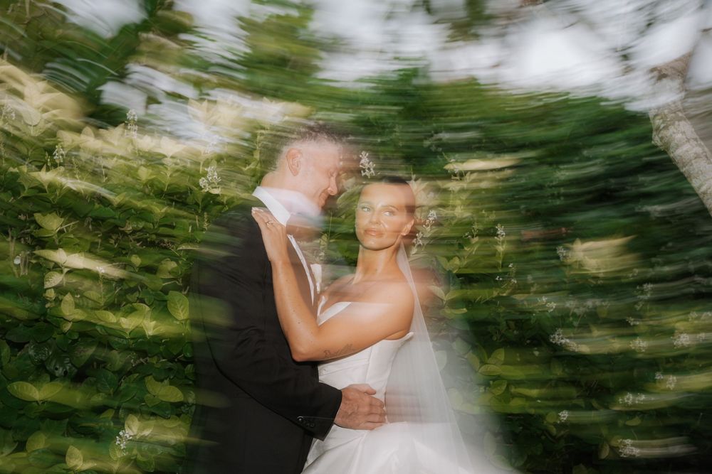 A wedding couple embraces while standing in front of a blurred green forest background.