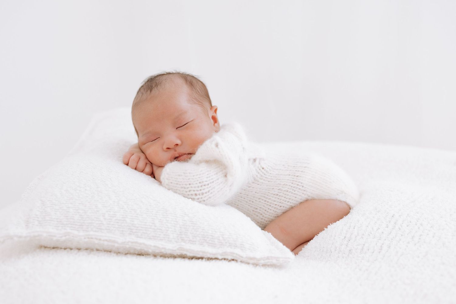 Peaceful sleeping baby wearing white knit clothing rests on soft white bedding.