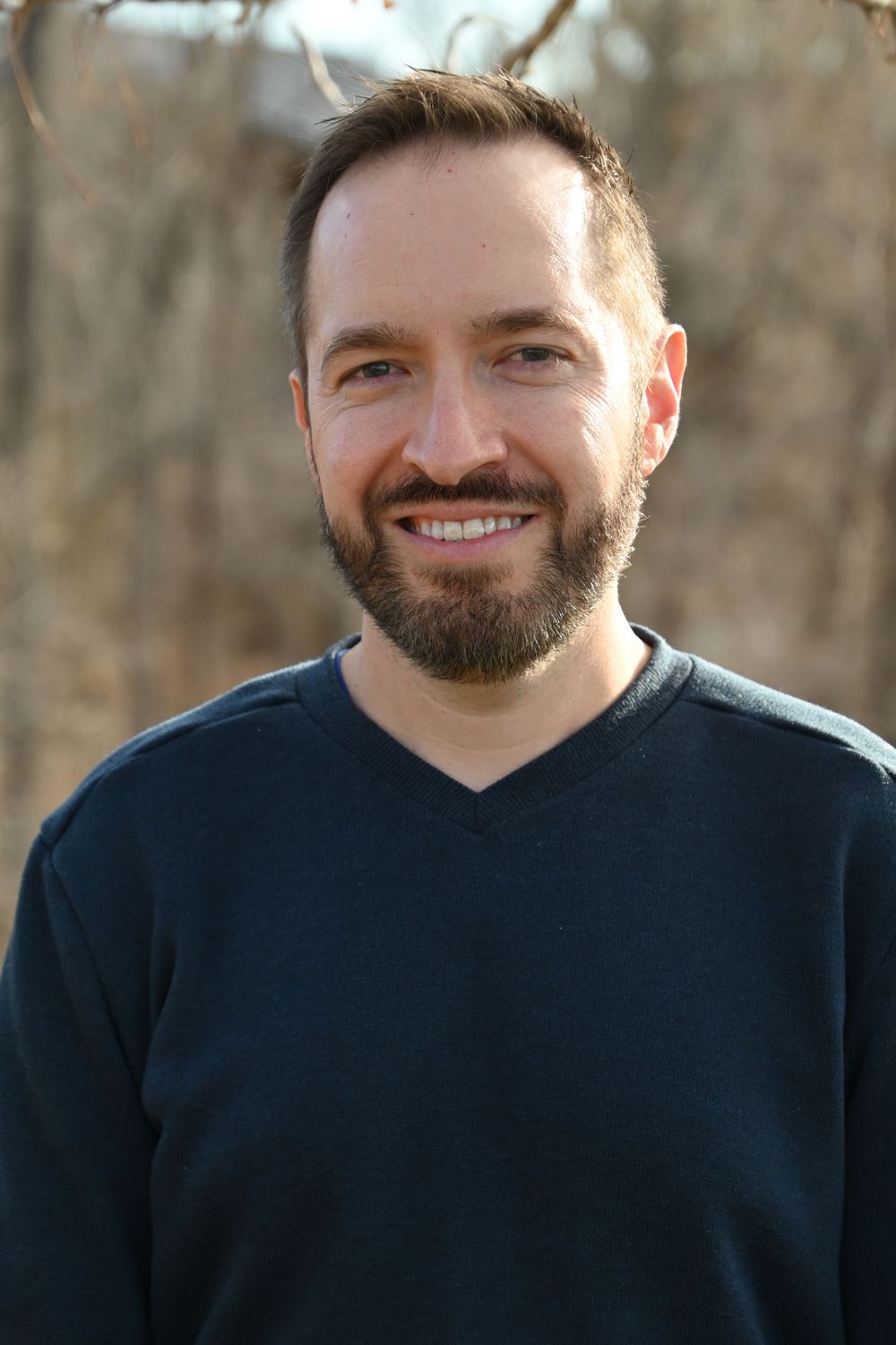 Professional headshot portrait of someone wearing a dark v-neck sweater smiling outdoors with blurred natural background.