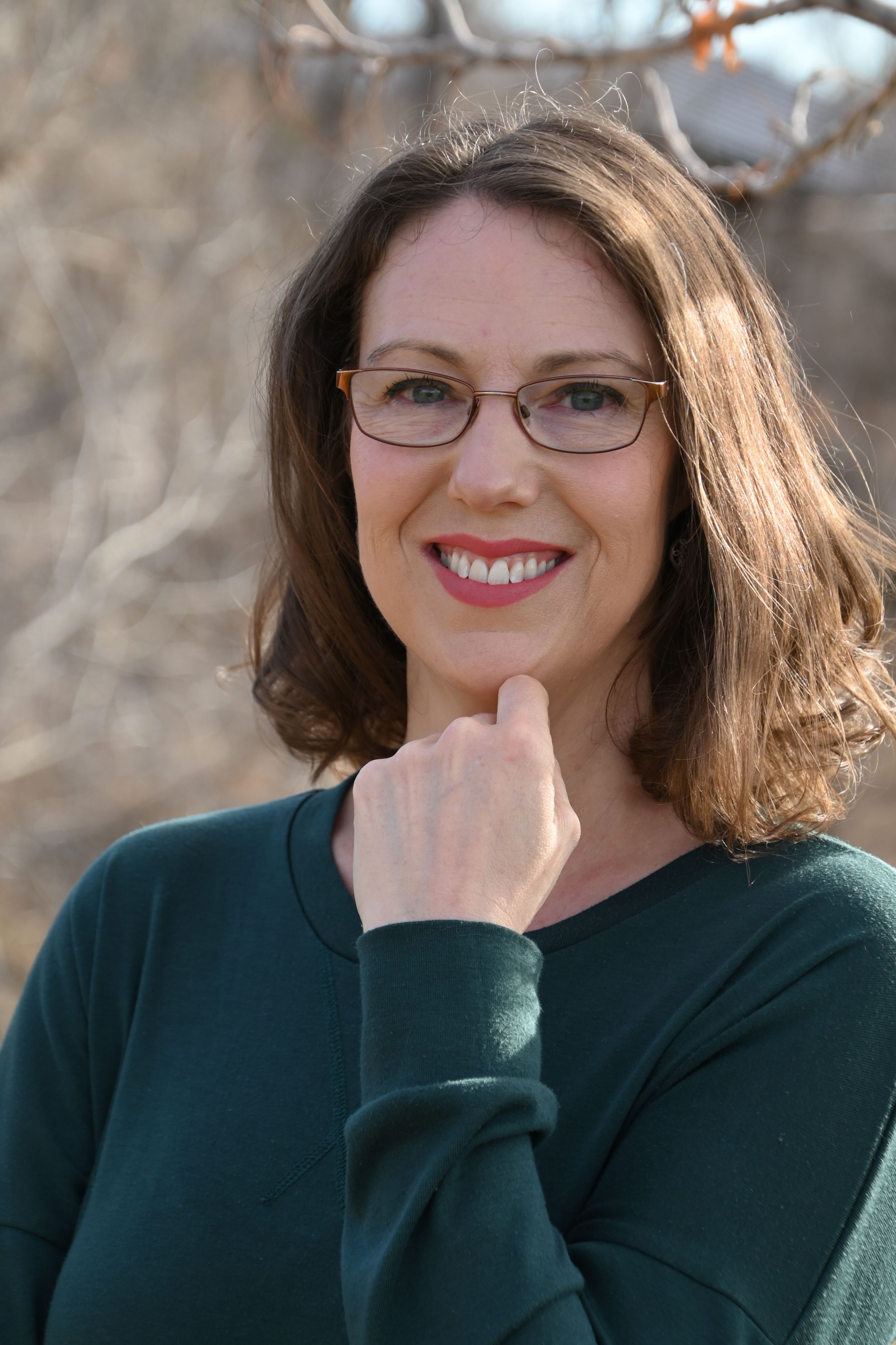 Professional headshot portrait with shoulder-length brown hair, glasses and dark green sweater against blurred outdoor background.