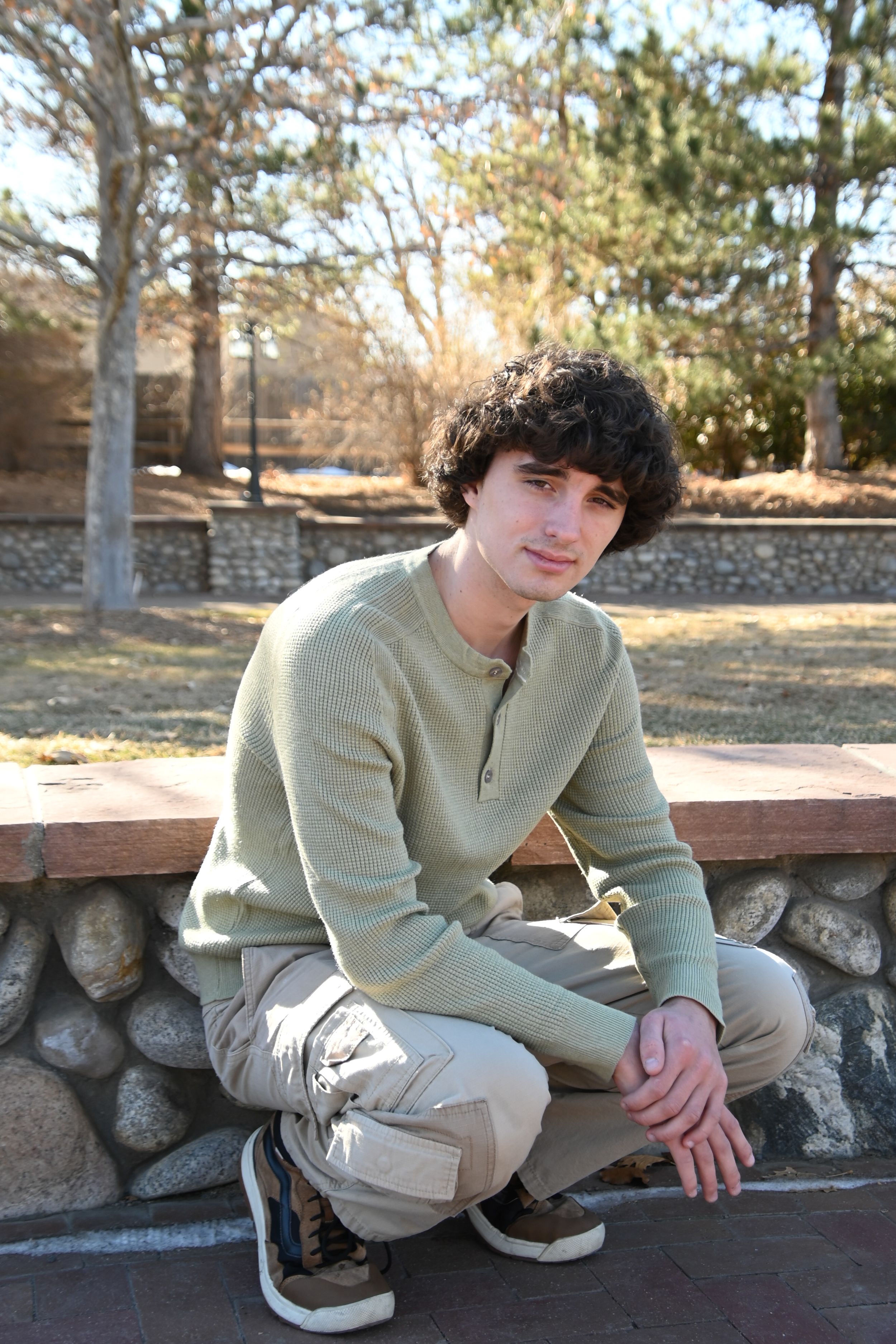 Someone in a light sage green sweater and khaki pants sits on a stone wall on a sunny autumn day.
