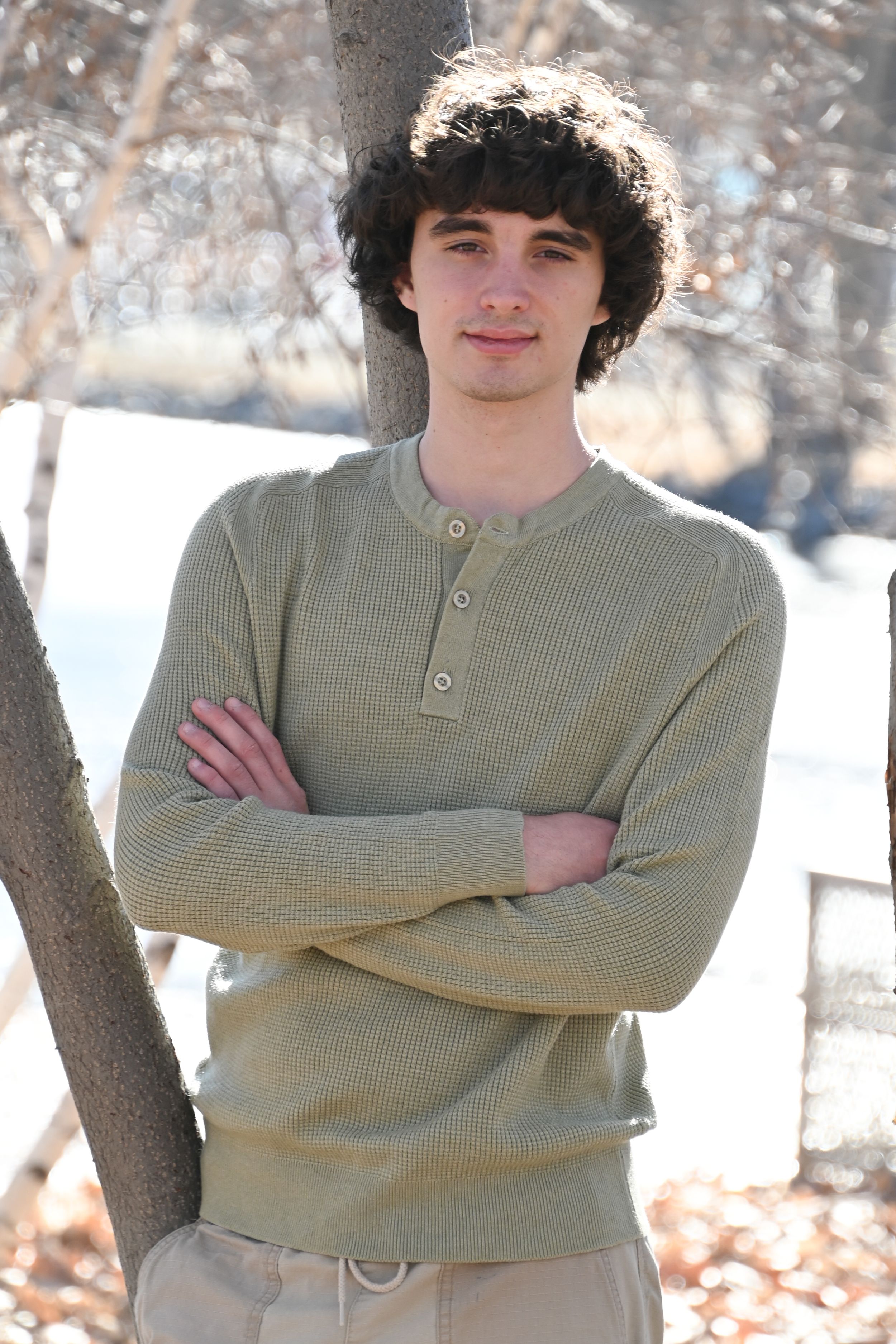 A person in a beige henley sweater stands with crossed arms against a tree in a snowy winter setting.