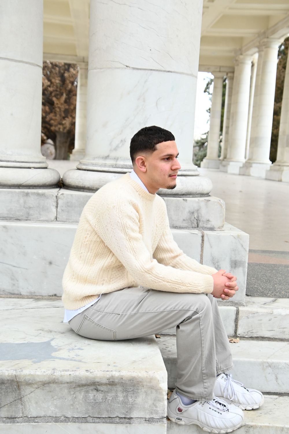 Someone in a cream cable knit sweater and grey pants sits on white marble steps near classical columns.