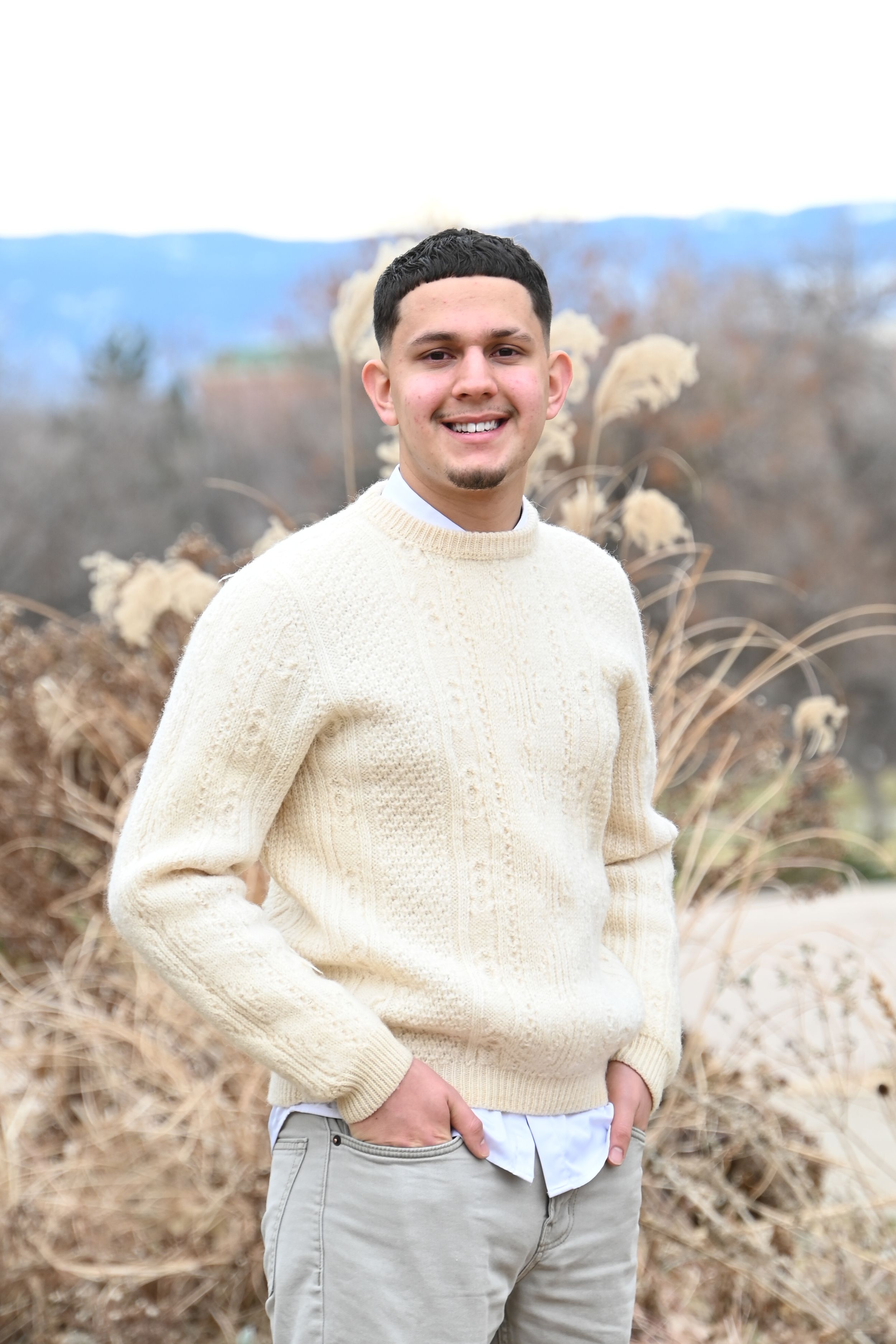 Person wearing beige cable knit sweater and light colored pants poses outdoors in a natural field setting.