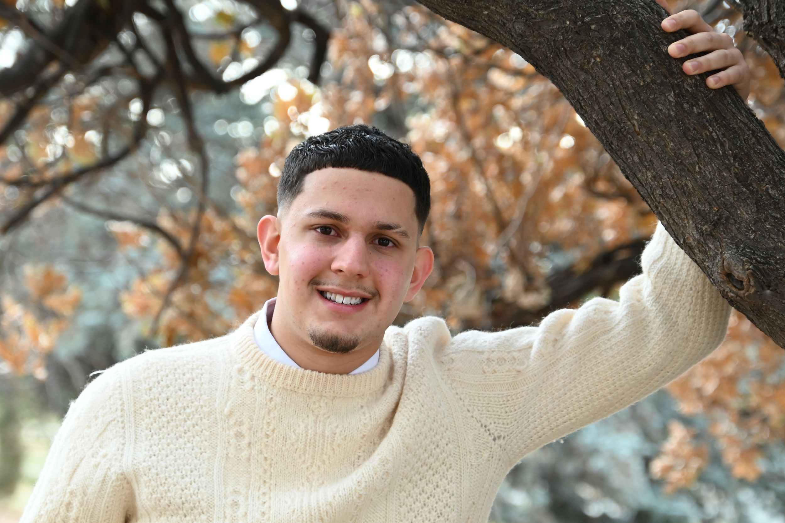 A person in a cream knit sweater poses playfully in autumn woods with brown leaves and trees.