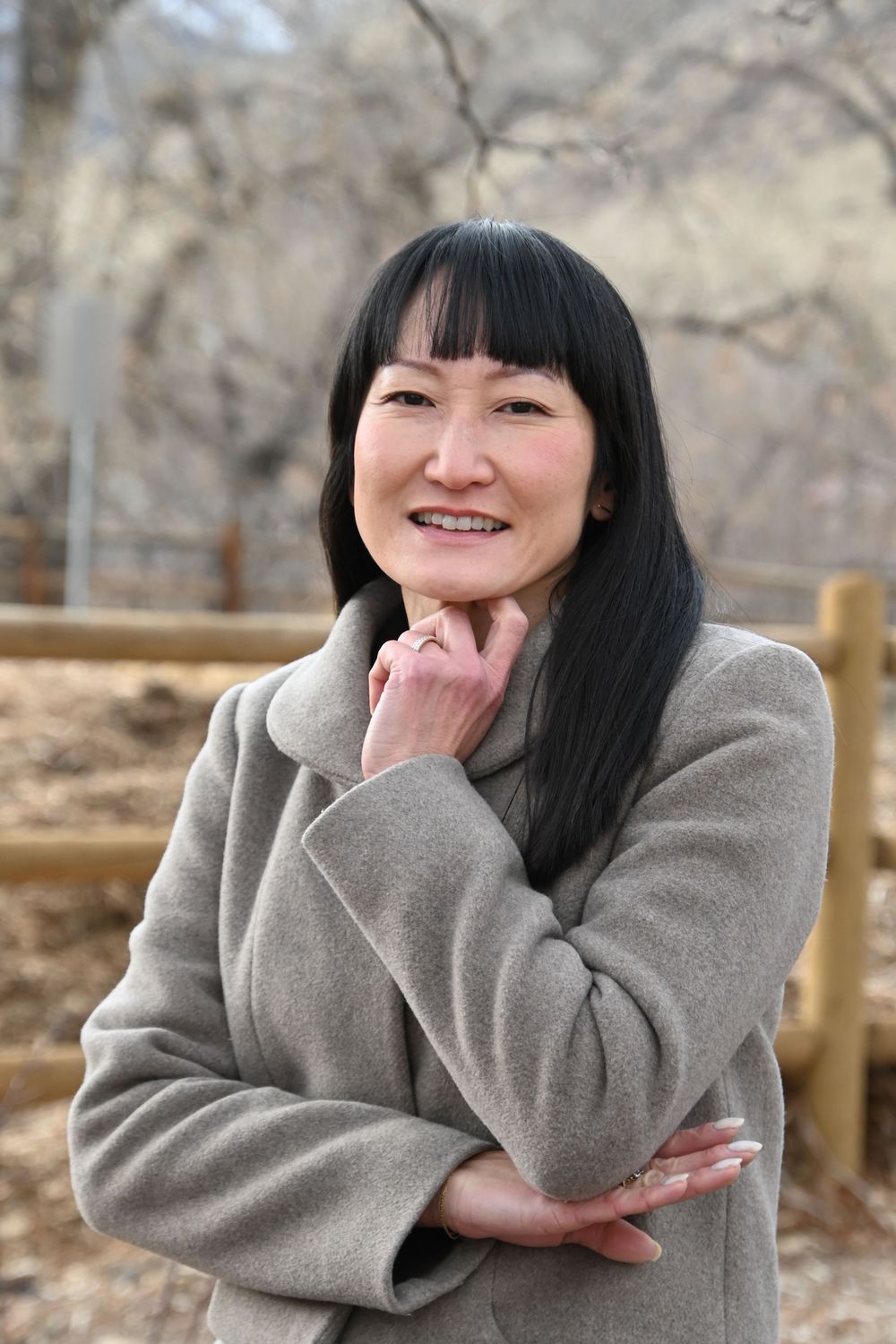 Smiling woman in gray winter coat poses outdoors with hand under chin against blurred natural background.
