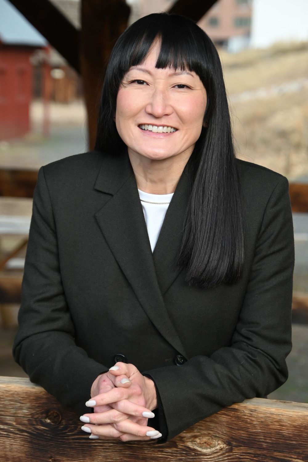 Professional portrait of a person with straight black hair wearing a dark blazer sitting at a wooden table.
