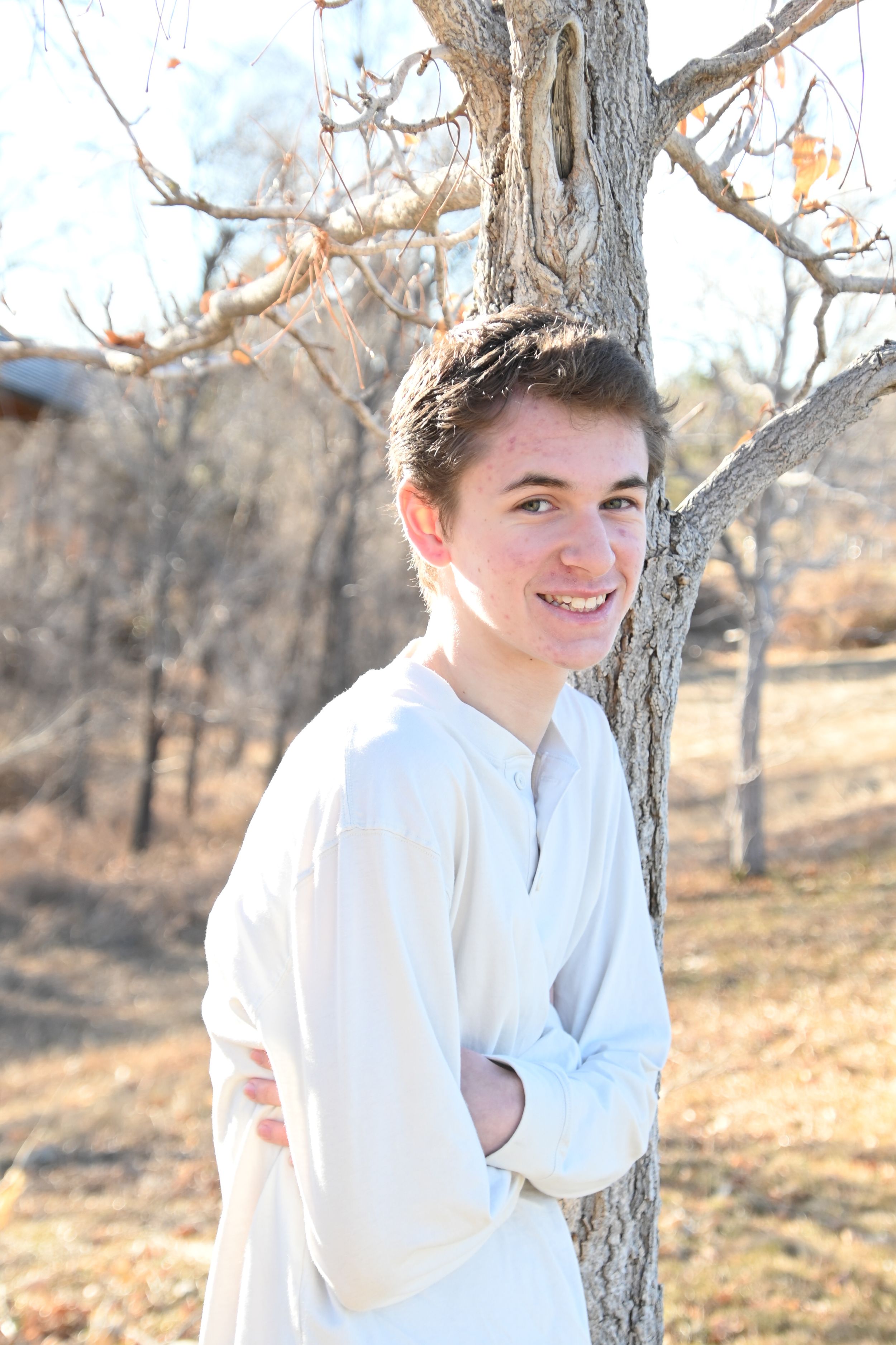 Portrait of someone in a white shirt leaning against a tree trunk in a wooded autumn setting.