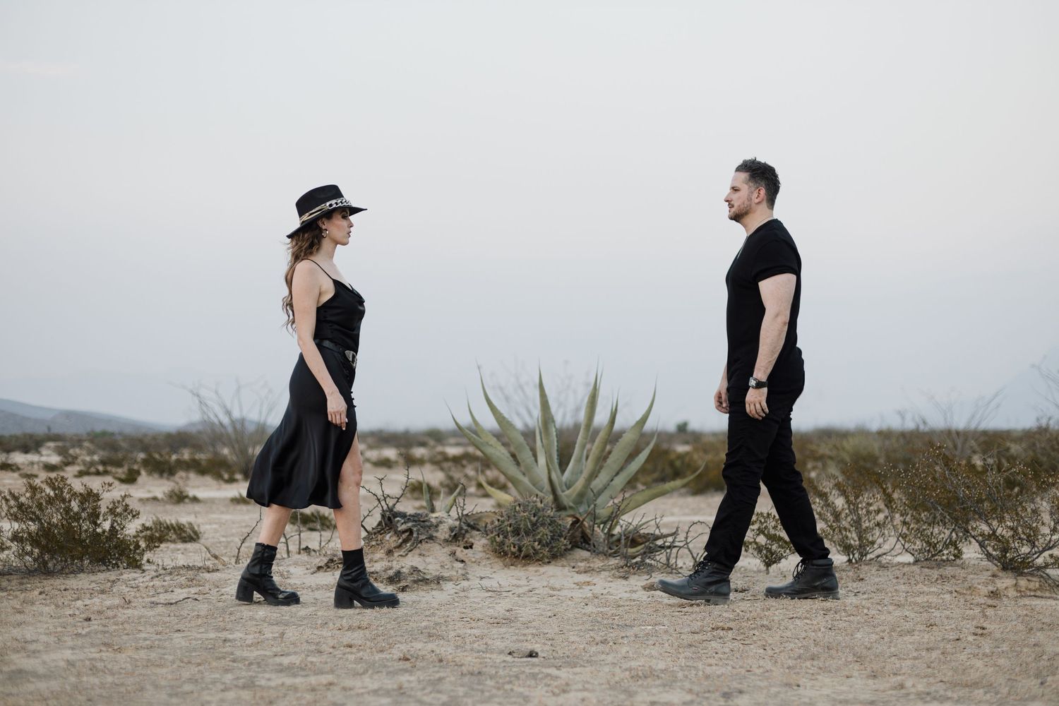 Couple in black outfits walking through desert landscape with agave plant.