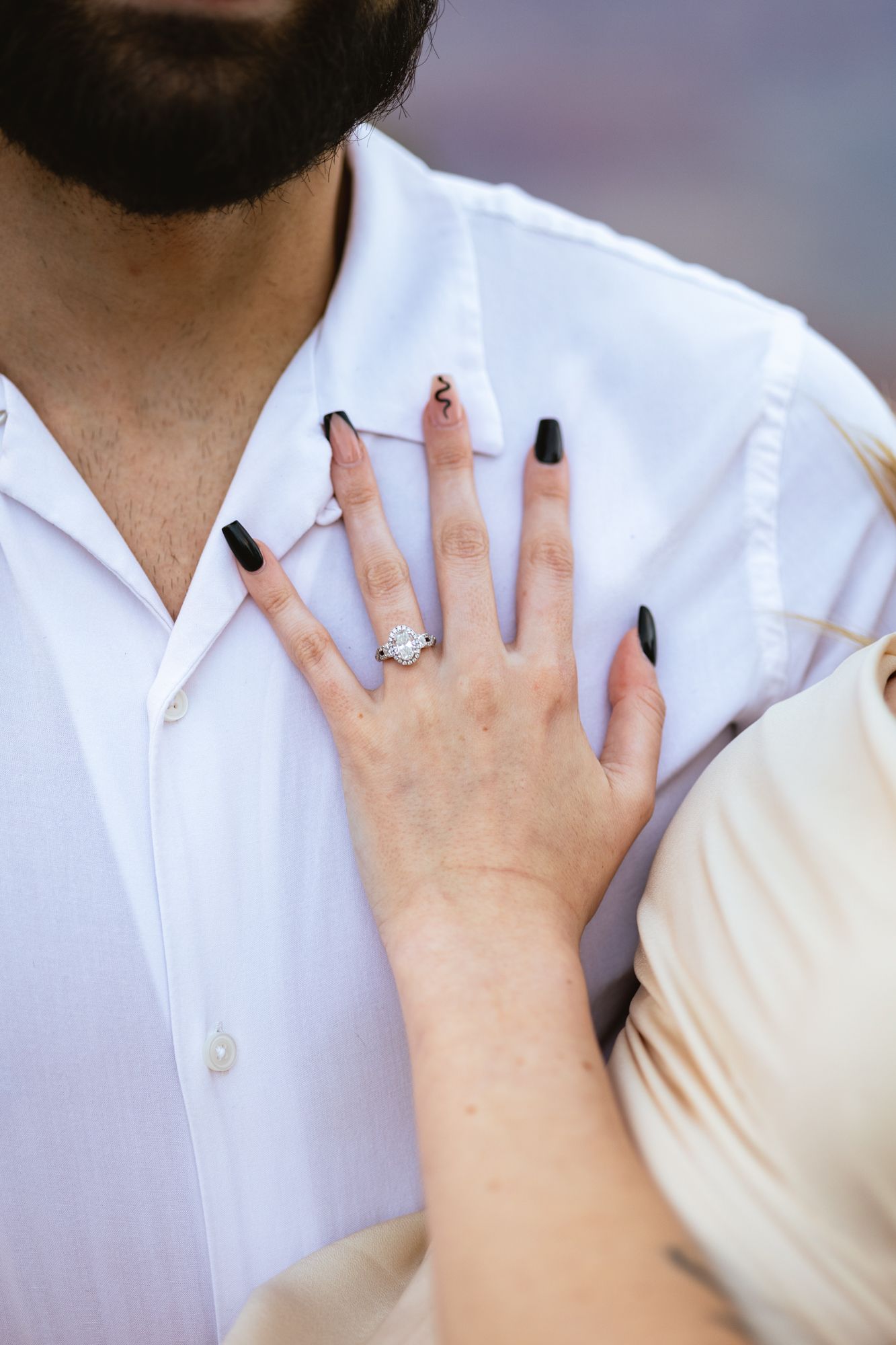 A woman's hand with black nail polish resting on a white collared shirt displays an engagement ring.