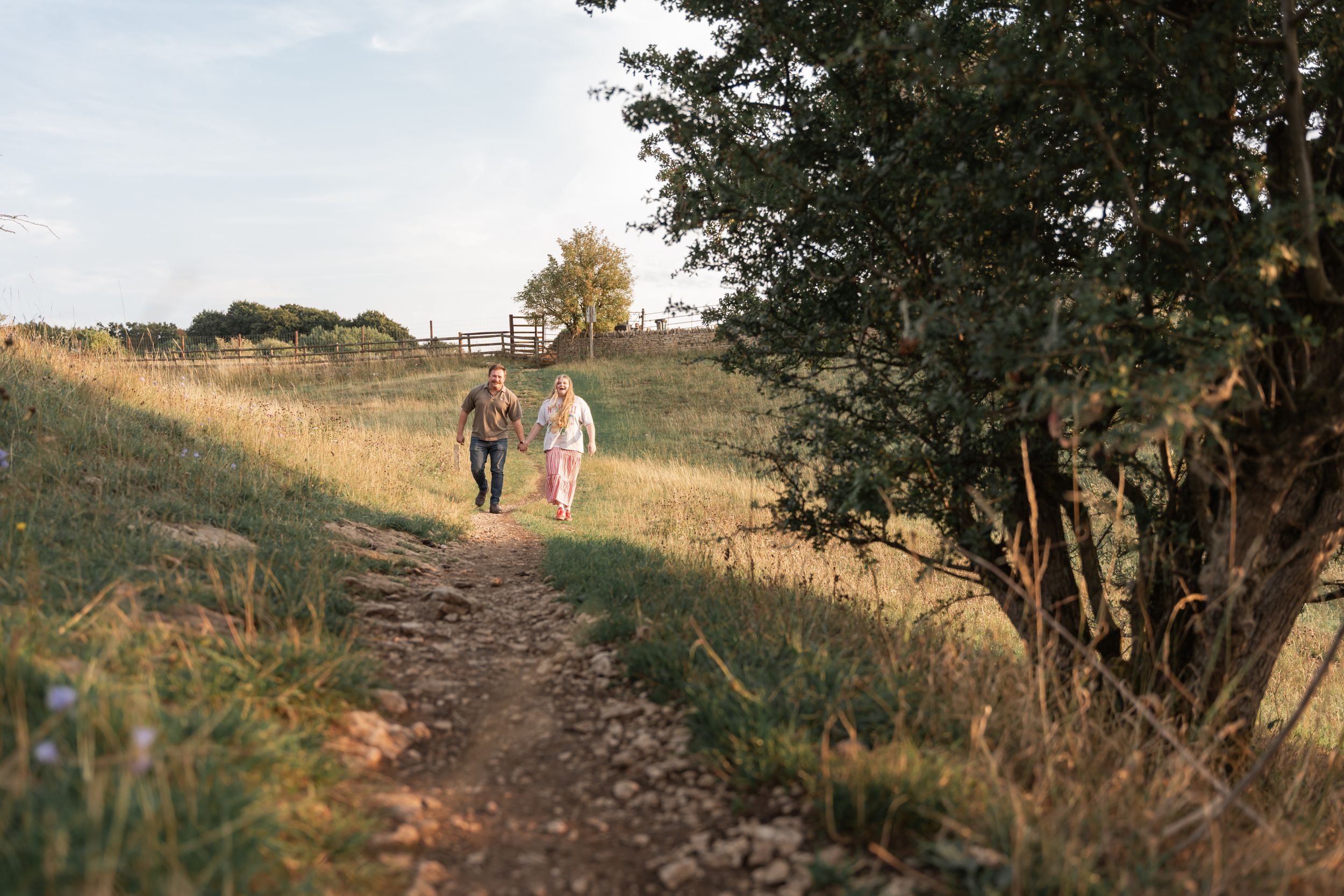 Two people walk together on a winding dirt path through golden grass at sunset with trees in the background.