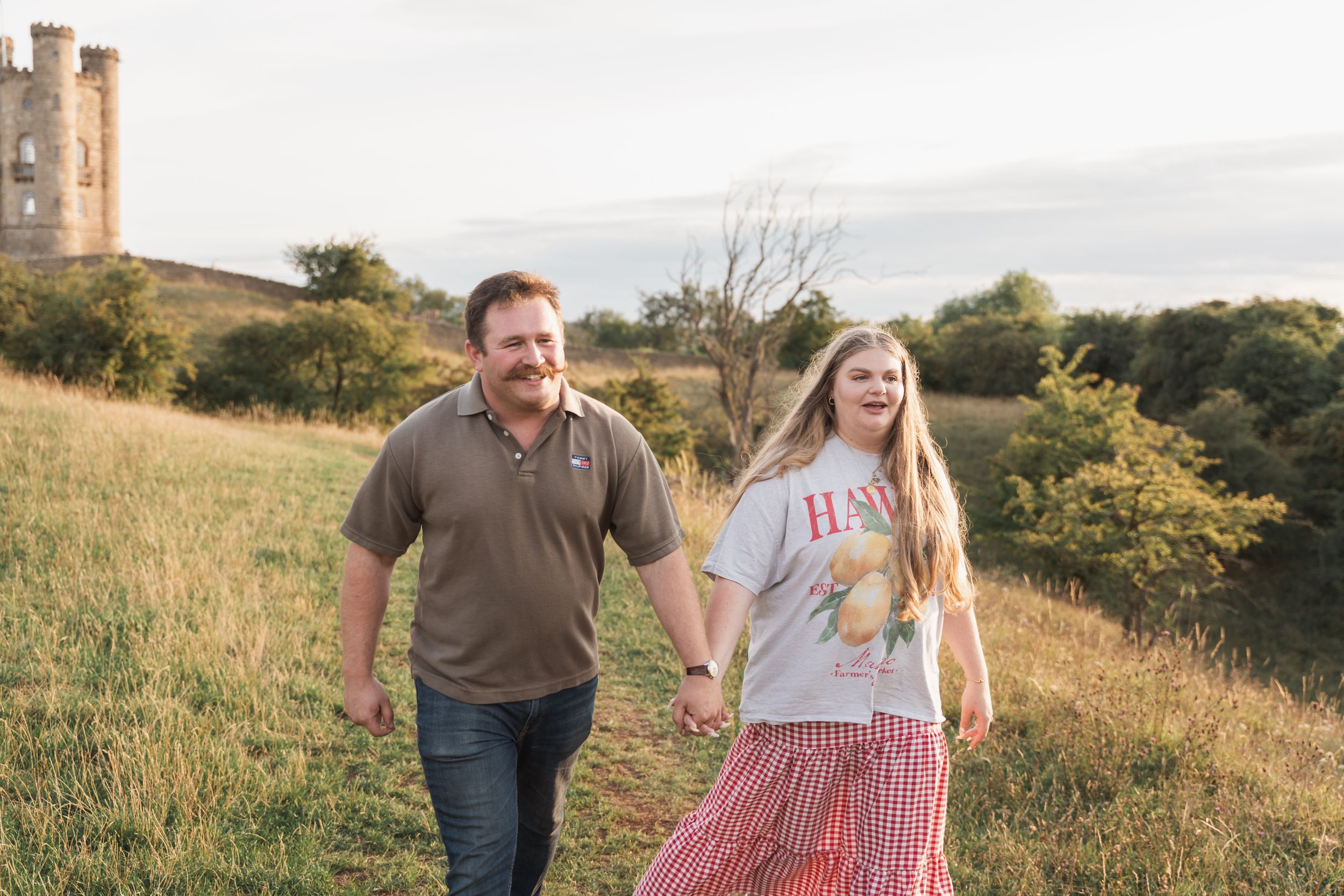 Two people take an evening stroll together up a grassy hill path with an old castle tower visible in the background.