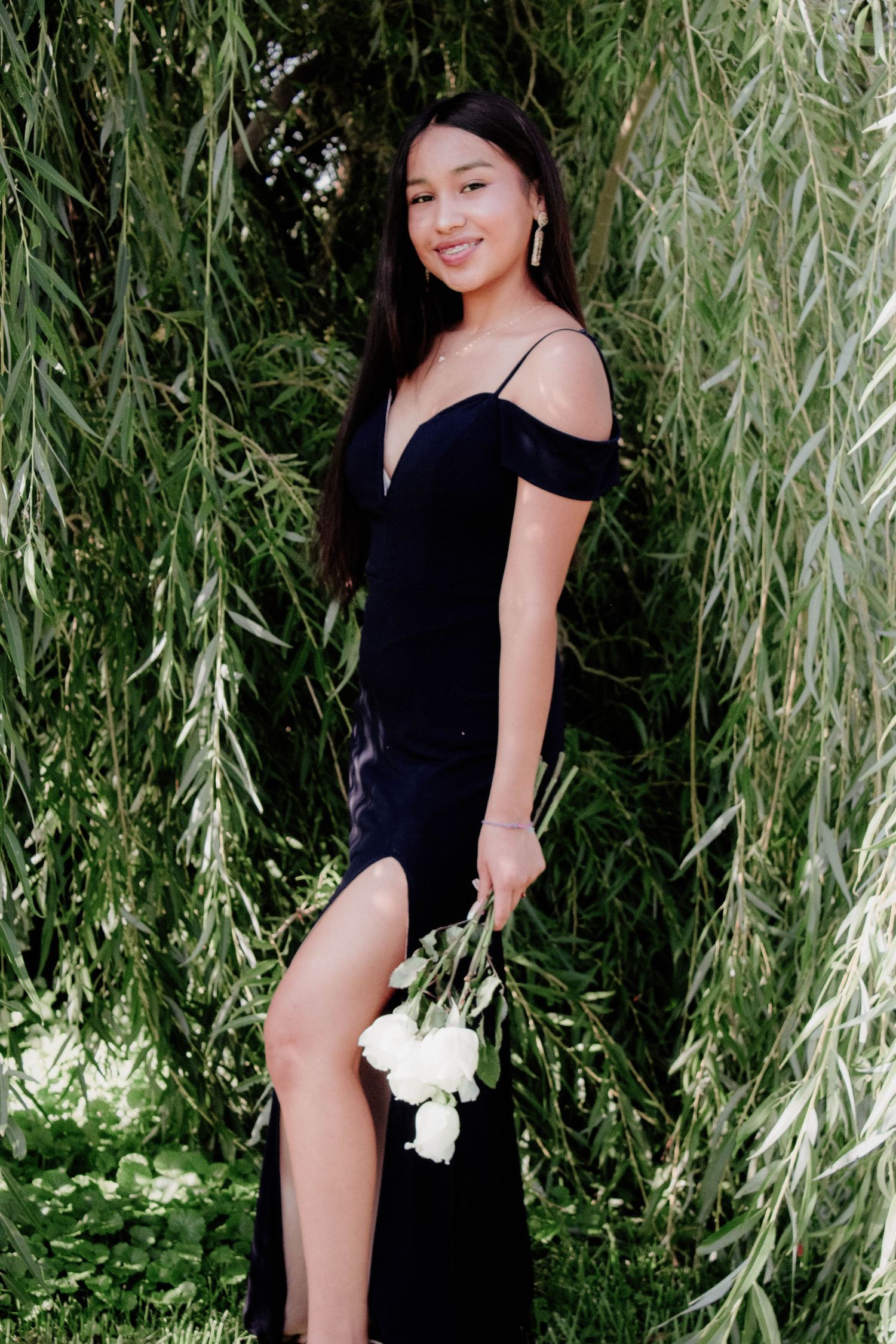 Elegant black formal dress with off-shoulder design and side slit photographed against cascading willow branches.