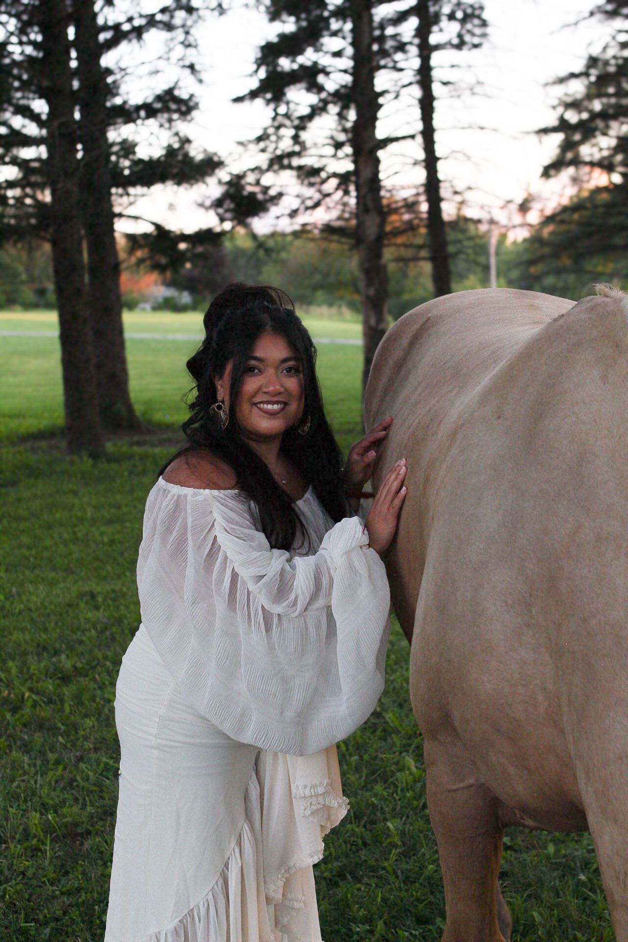 Person in flowing white dress stands beside light-colored horse in wooded area.
