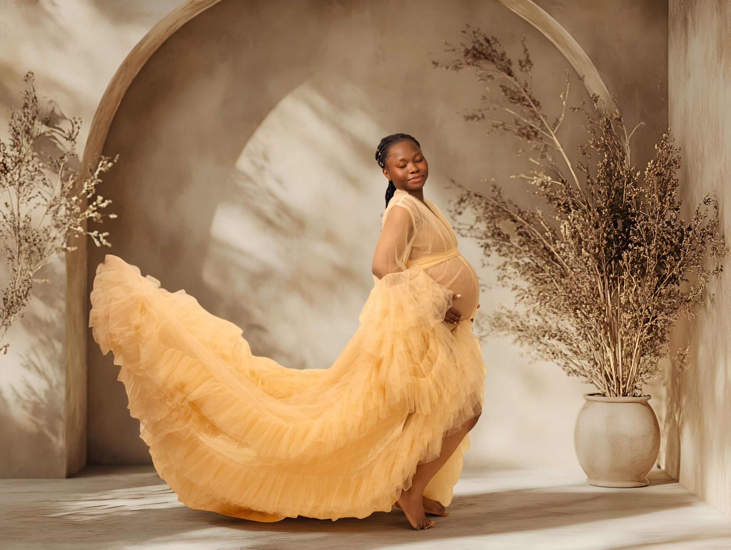 Pregnant woman in flowing yellow tulle gown poses gracefully in elegant neutral studio setting with dried botanicals.