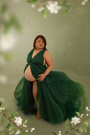 Pregnant woman in elegant emerald green tulle gown posing gracefully among white flowers on a soft green background.