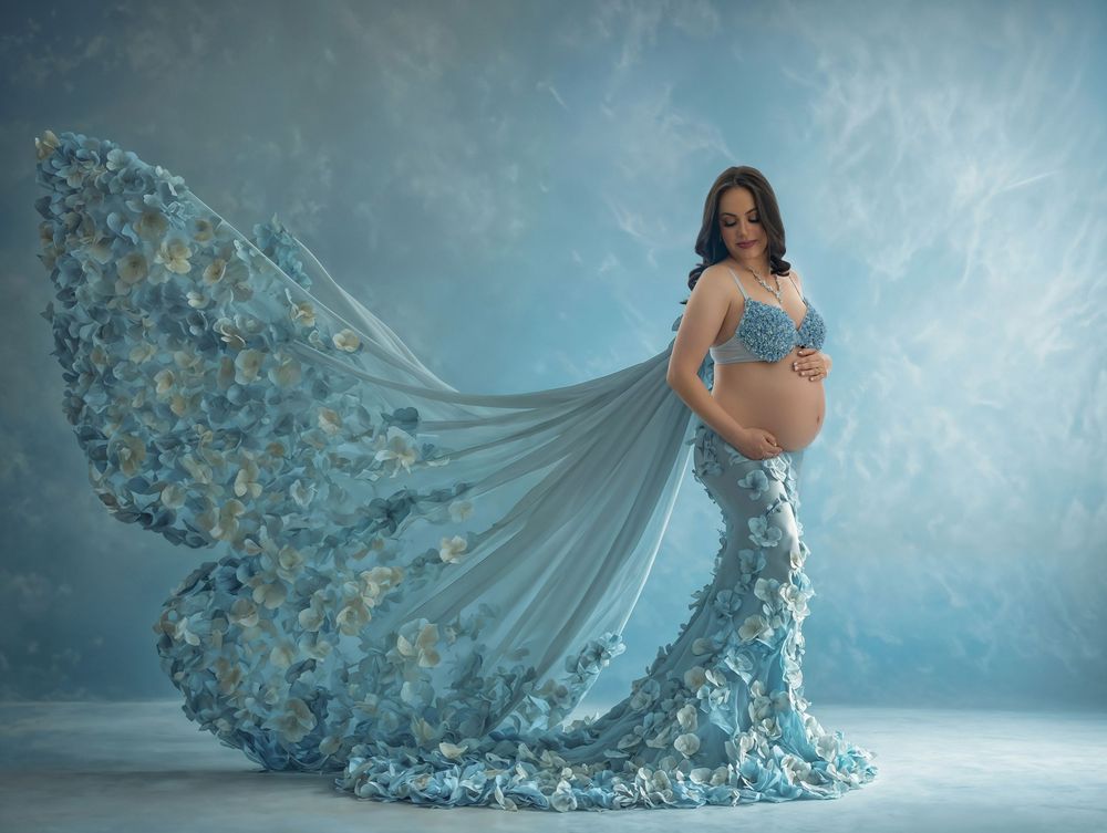 A figure in a flowing blue couture gown with floral embellishments poses dramatically in an ethereal studio setting.