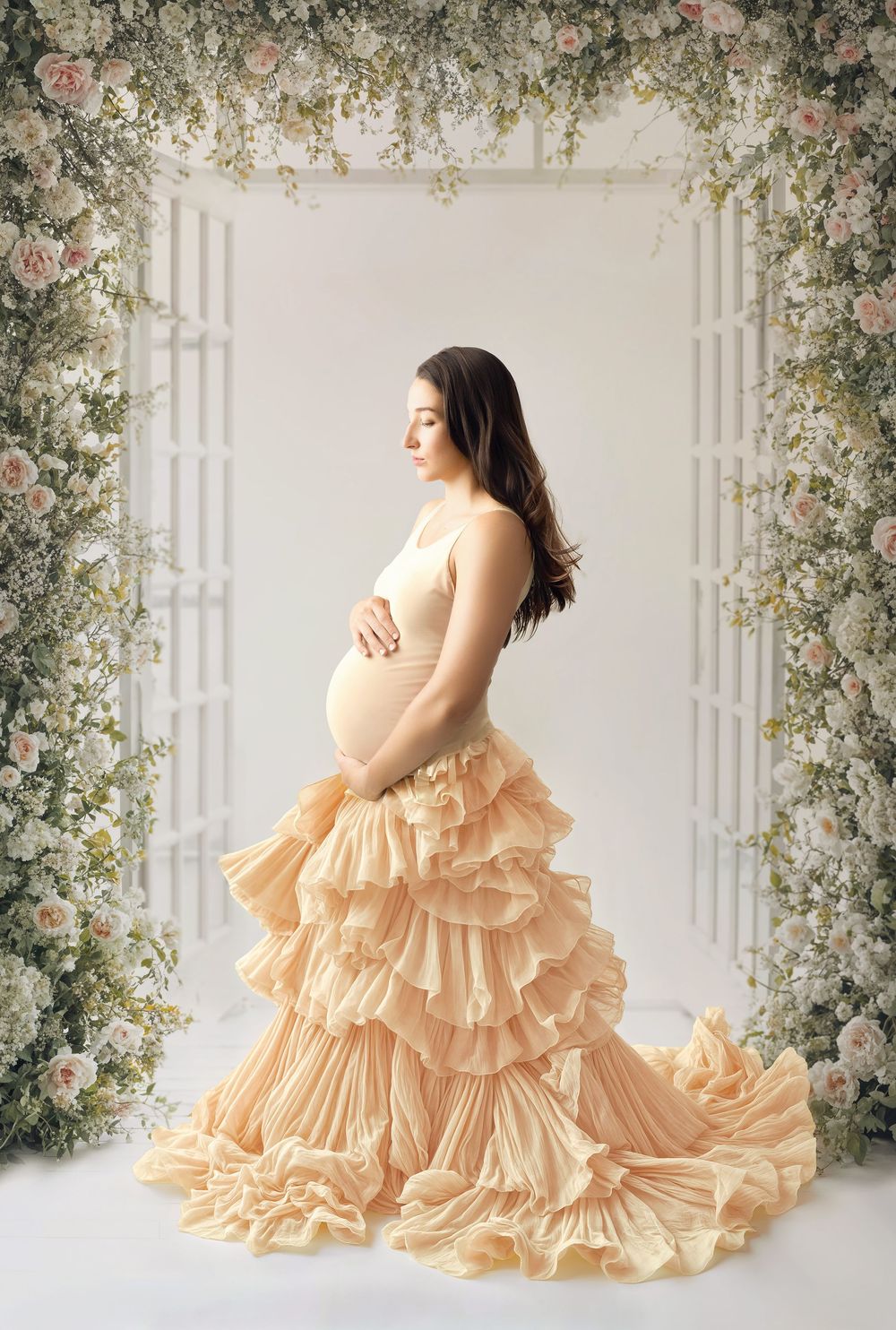 A maternity portrait featuring a flowing peach ruffled gown against a romantic floral arch backdrop in soft lighting.