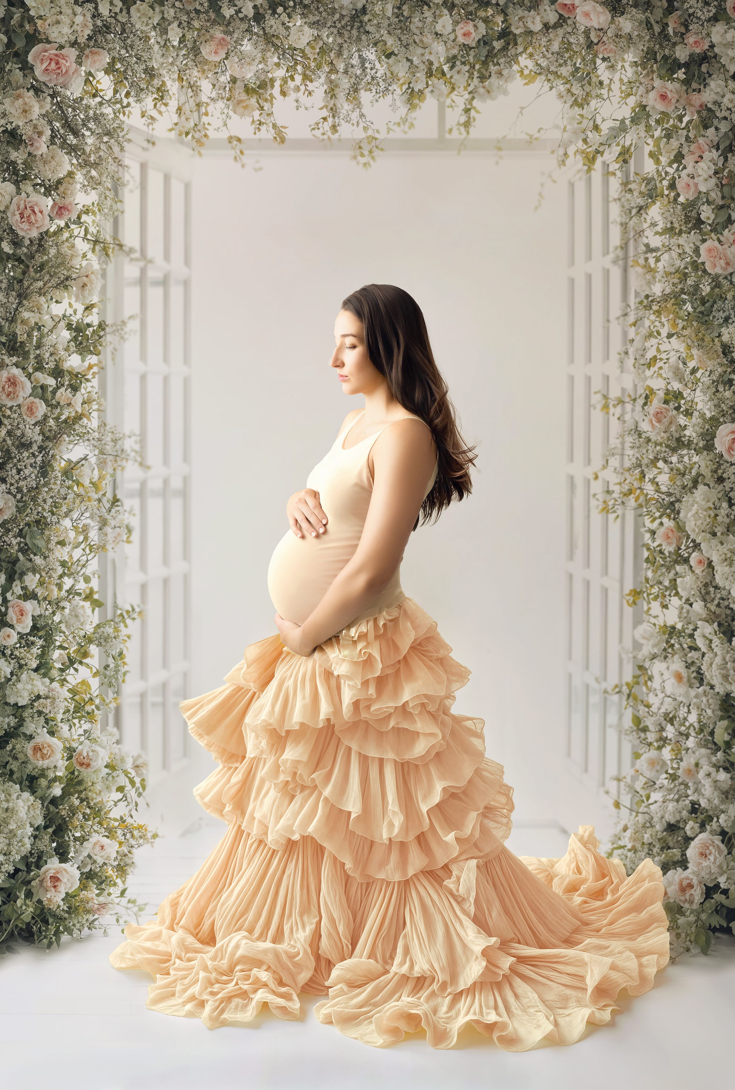 A maternity portrait featuring a flowing peach ruffled gown against a romantic floral arch backdrop in soft lighting.